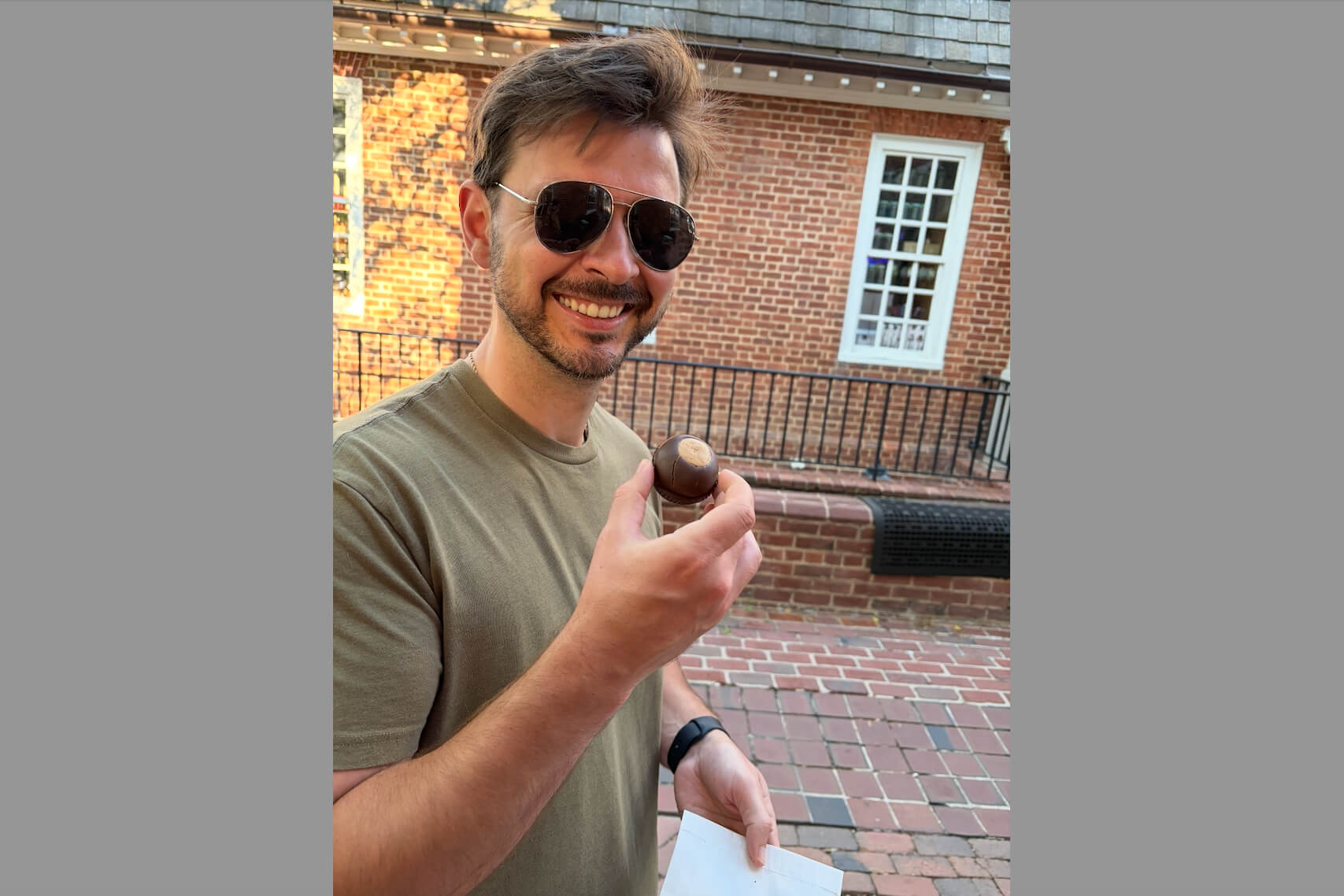 Man wearing sunglasses smiles while holding a round chocolate confection outdoors.