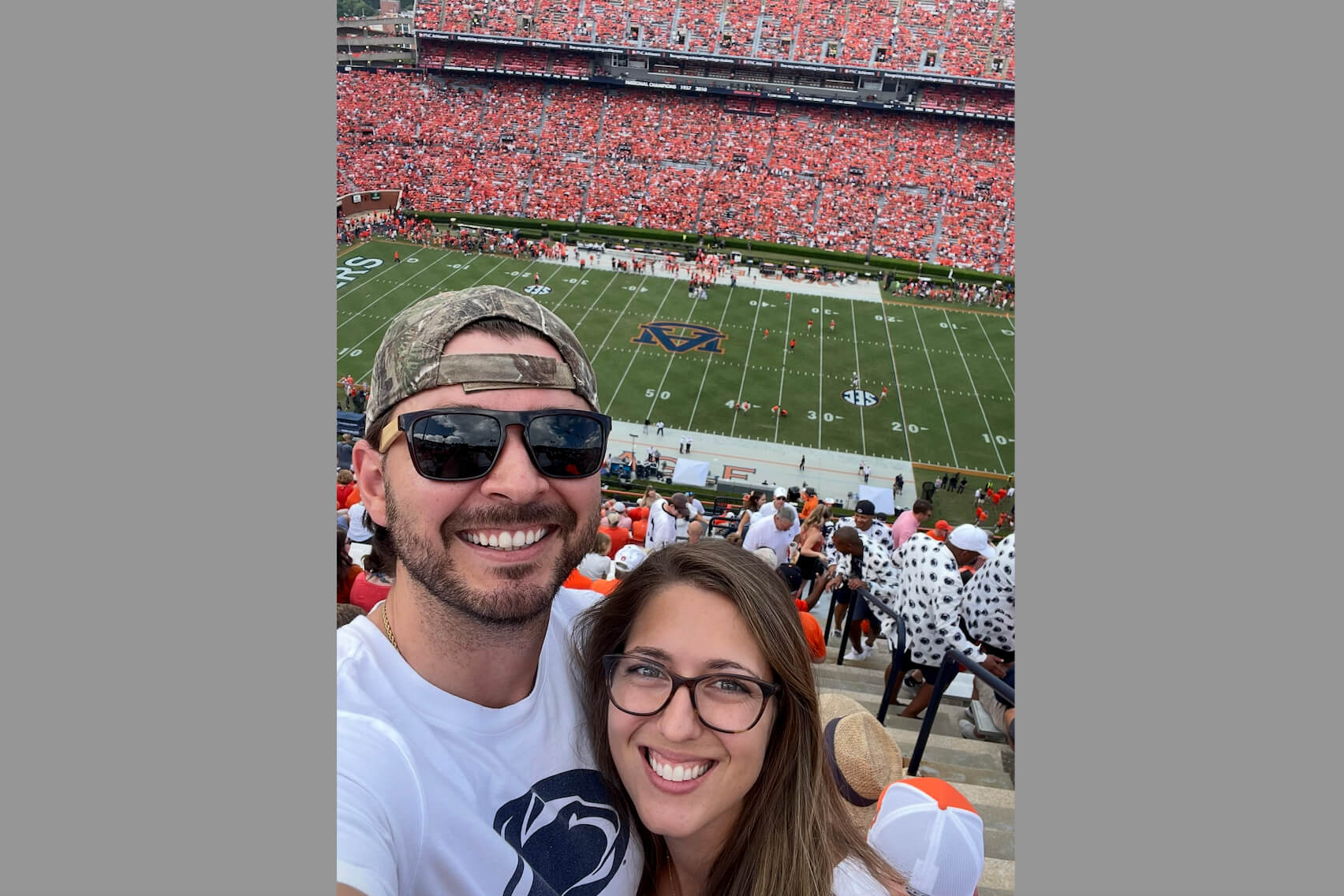 Smiling man and woman taking a selfie in a football stadium with a large red-clad crowd and a green field ahead.