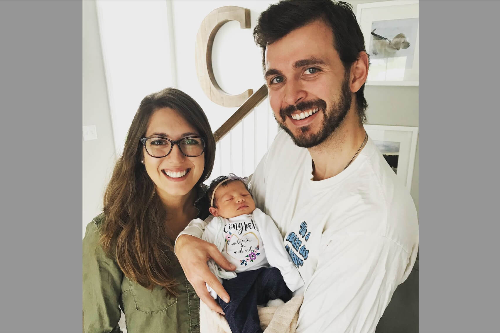 Smiling couple holds a newborn baby indoors near a staircase.