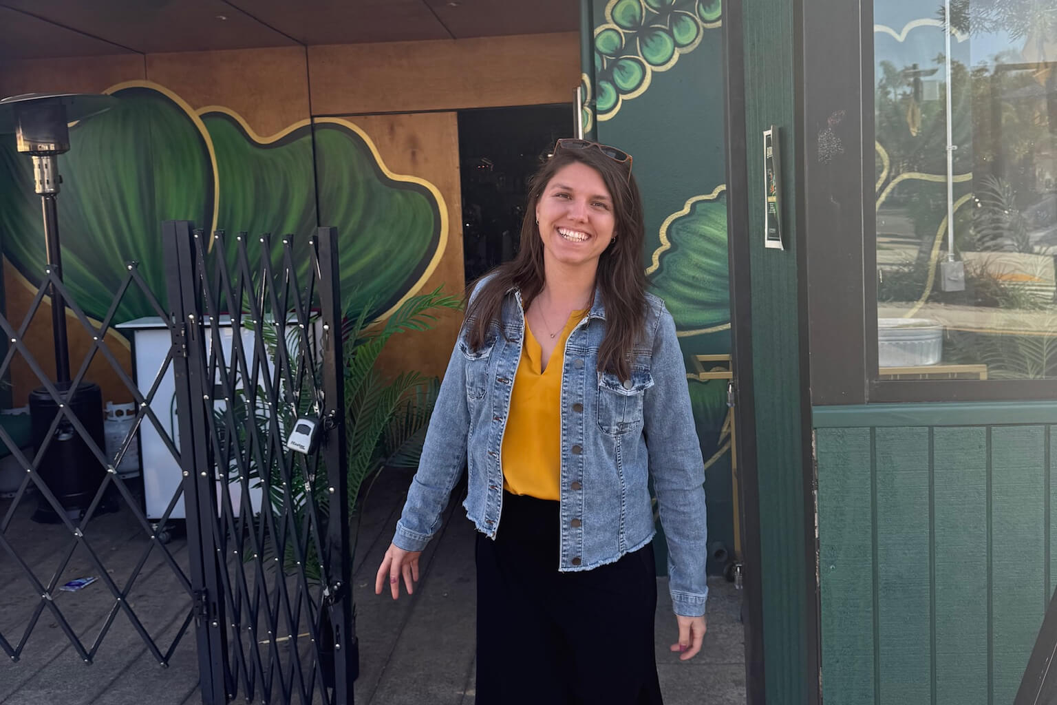 Smiling woman in a yellow blouse and denim jacket stands outside a muraled storefront, with a folded metal gate on her left.