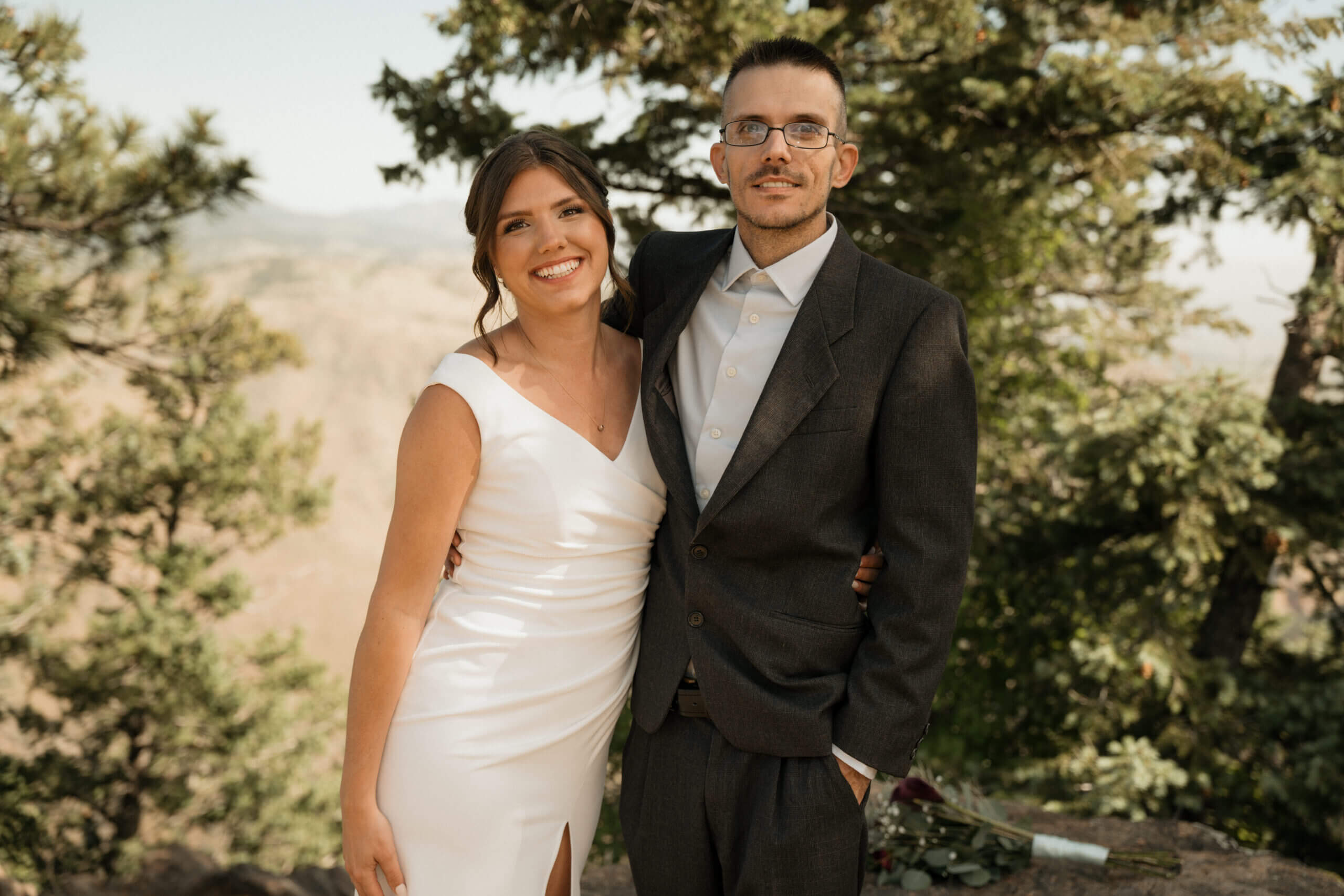 Smiling bride in a white dress and groom in a dark suit pose together outdoors among trees and greenery.