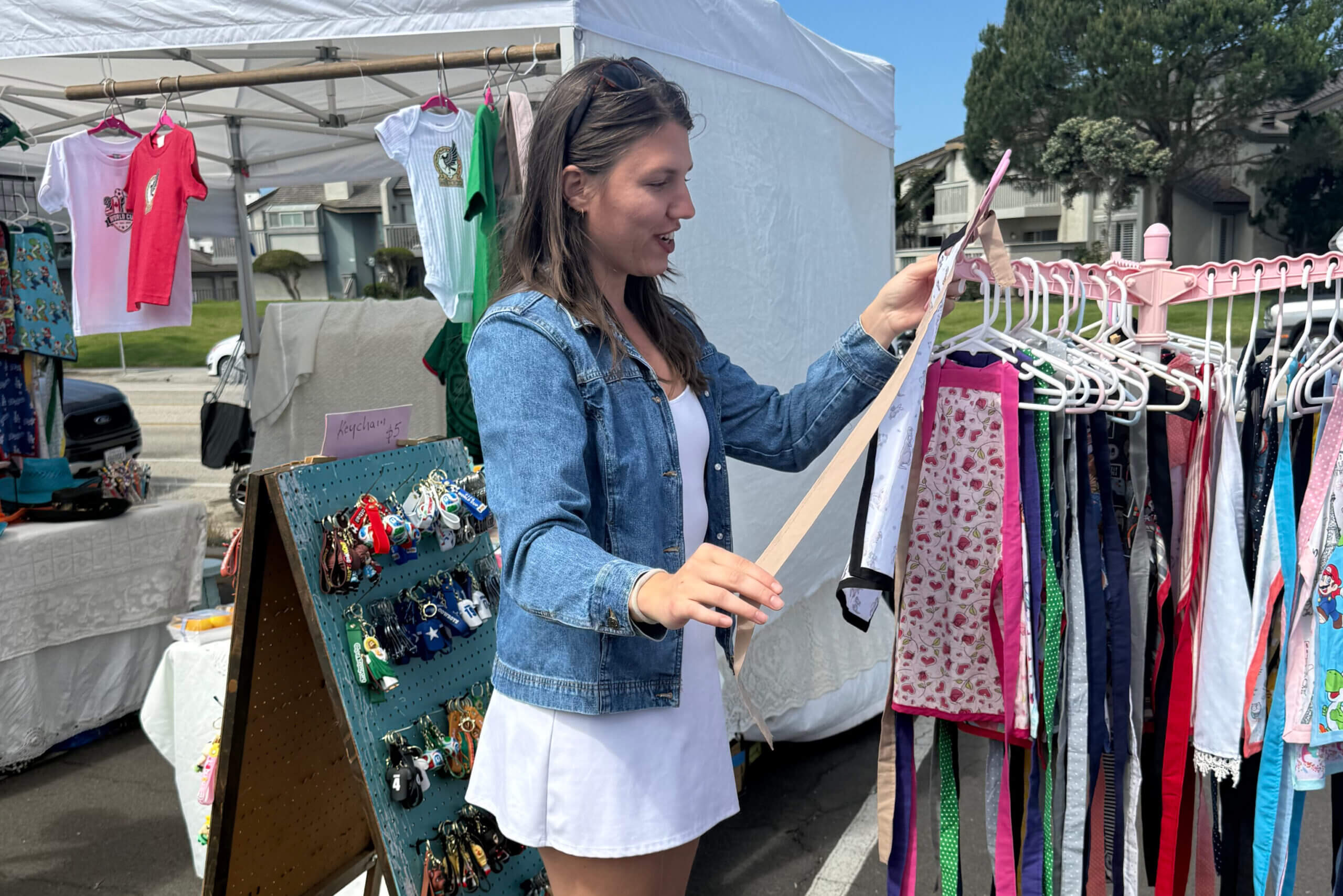 Woman in a denim jacket browsing colorful dresses at an outdoor market stall under a white canopy.
