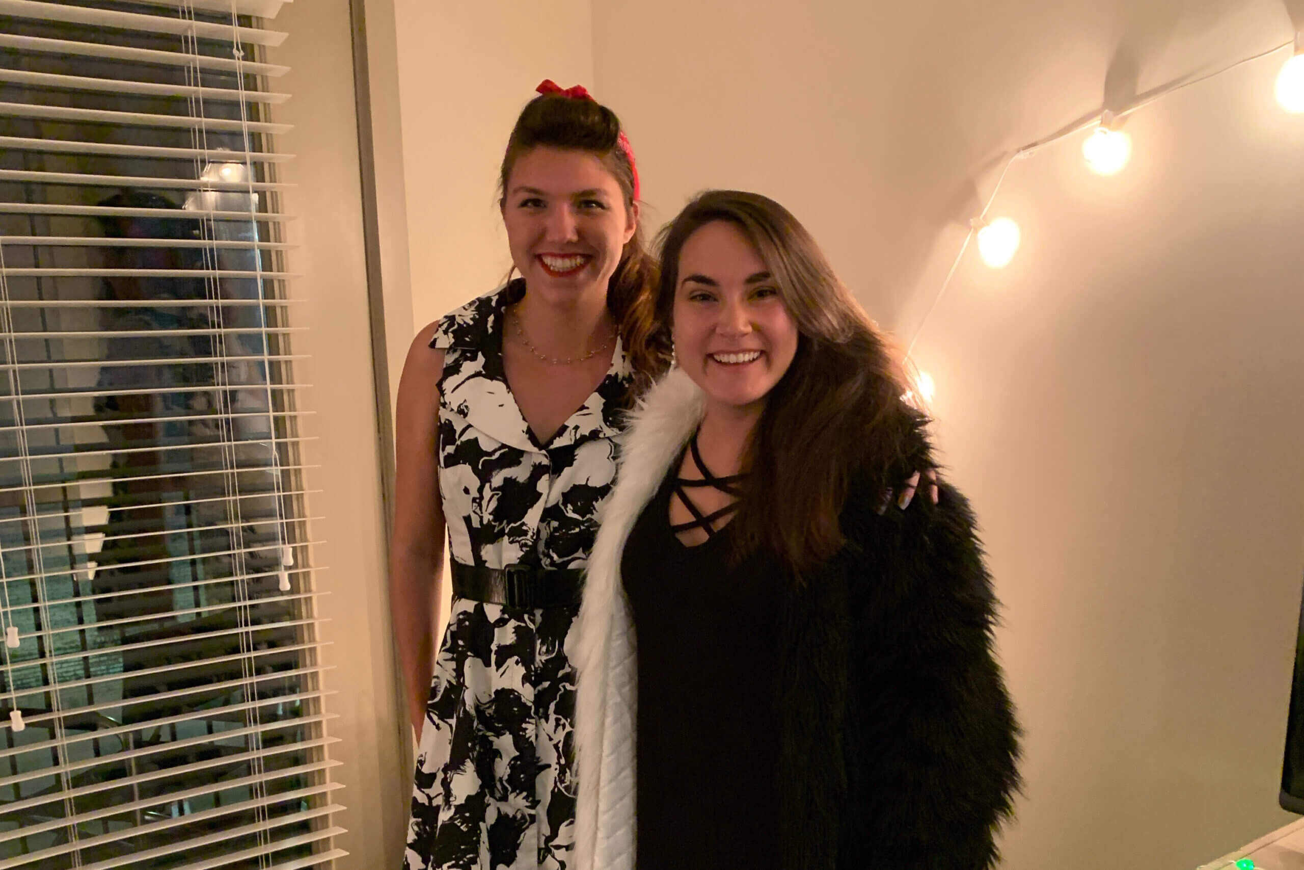 Two smiling women posing indoors, one in a black-and-white floral dress with a red headband and the other in black with a white fur stole, by string lights