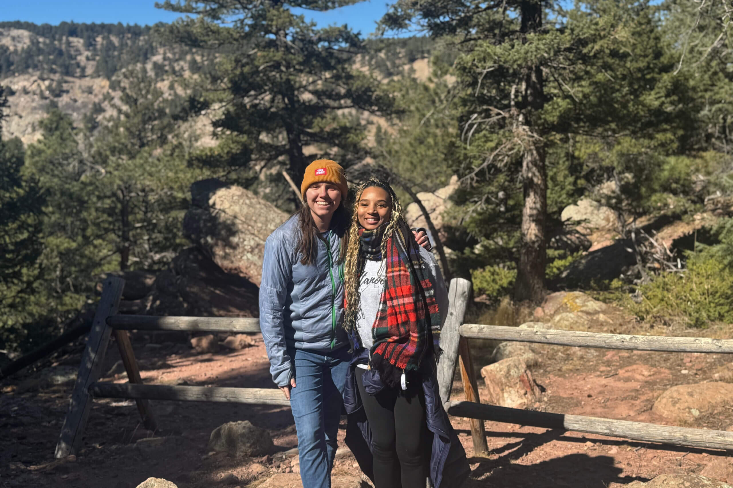 Two friends posing for a photo outdoors on a wooded hiking trail, smiling with a wooden railing in the foreground.