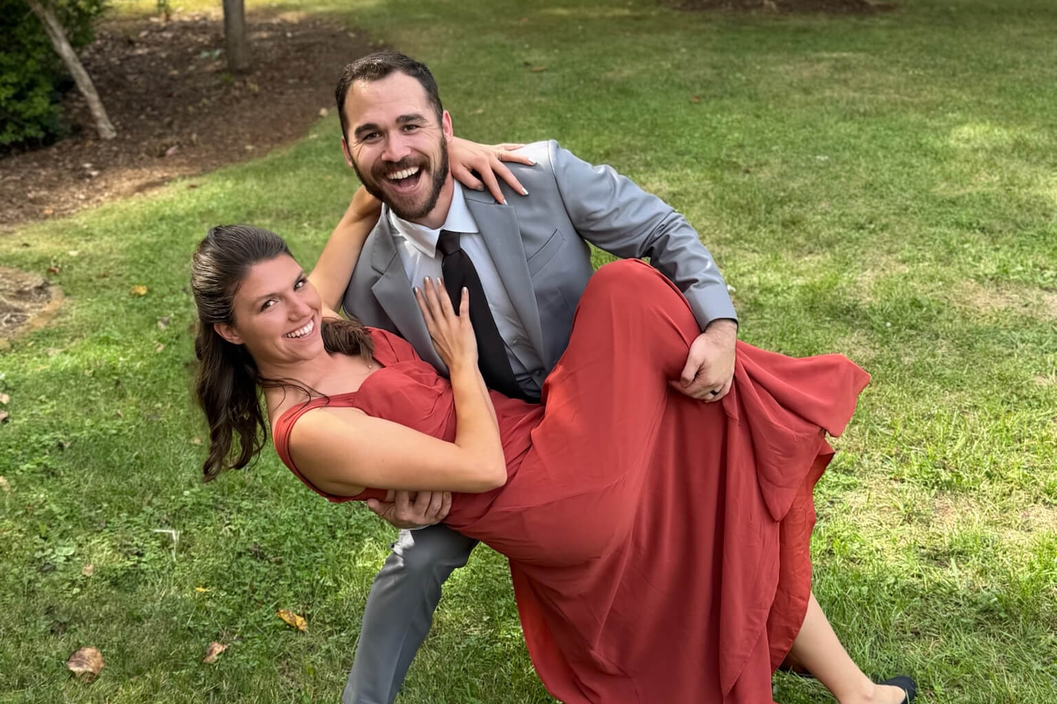 Smiling couple outdoors: man in gray suit lifts woman in red dress as they pose playfully on the grass.