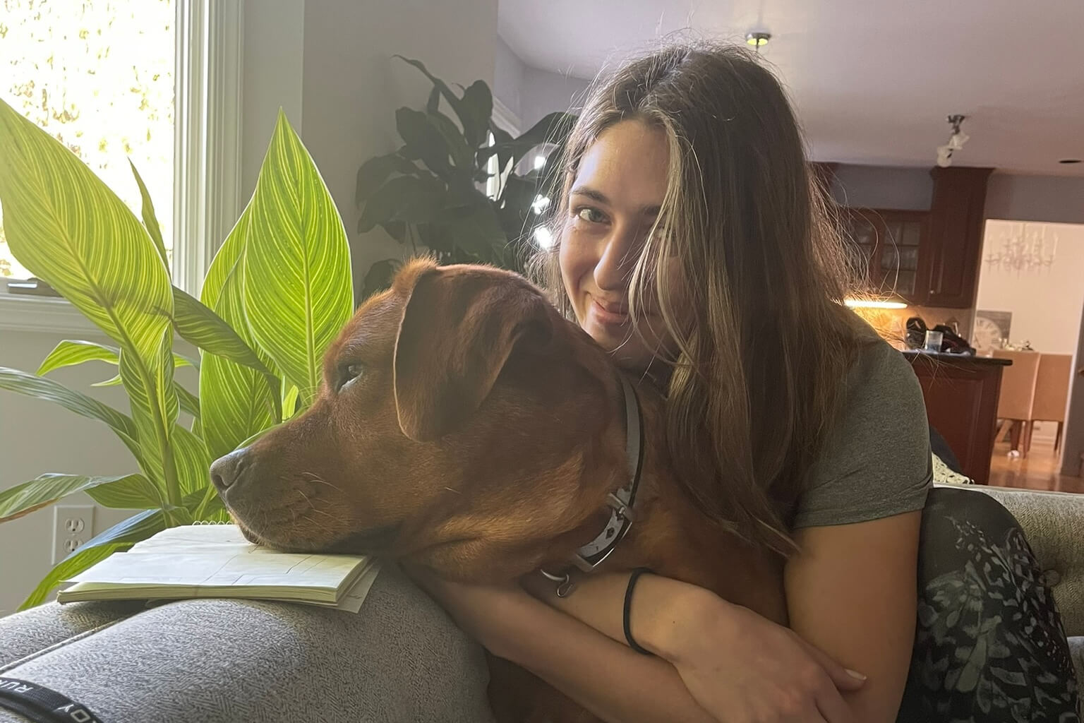 Young woman smiles while cuddling a brown dog on the couch near a notebook by a sunny window and a large green plant behind them.