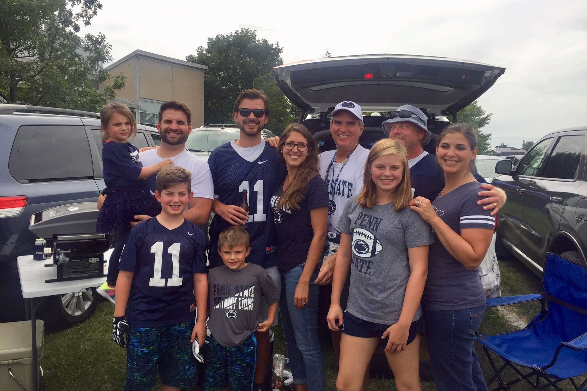 Group of Penn State fans, adults and children, posing for a photo at an outdoor tailgate by parked cars and a open trunk behind them.