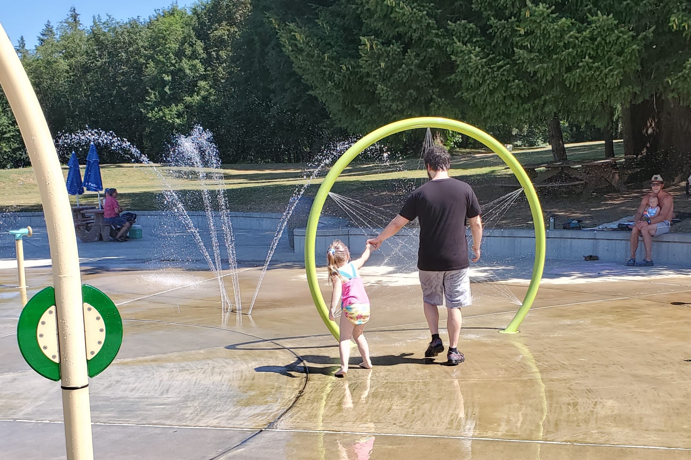 Father and young girl holding hands inside a bright lime-green circular water feature while jets spray nearby at a park splash pad: playful summer scene.