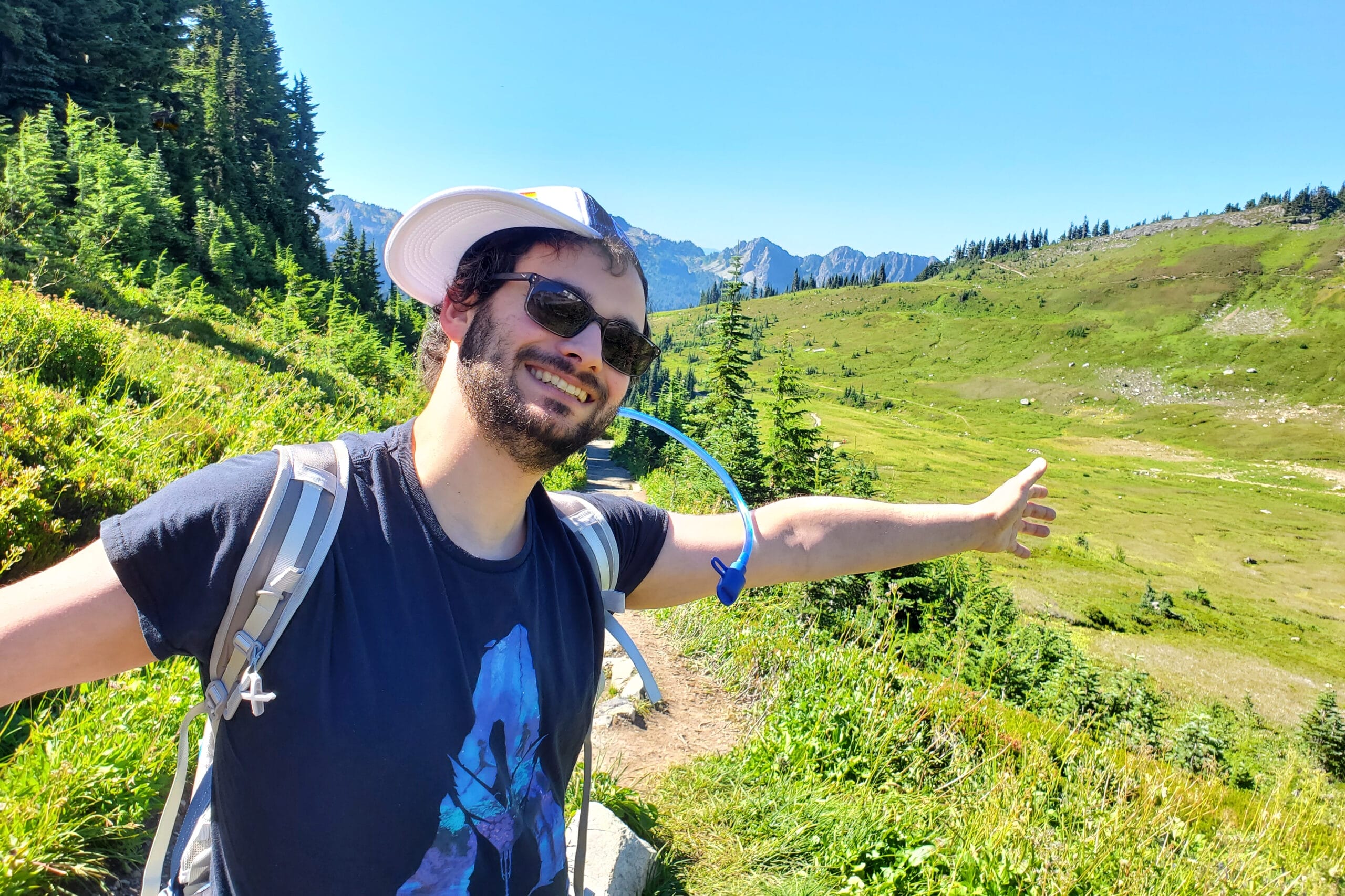 Smiling man wearing sunglasses and a cap, hiking backpack, arms outstretched on a mountain trail with green meadows and evergreen trees.