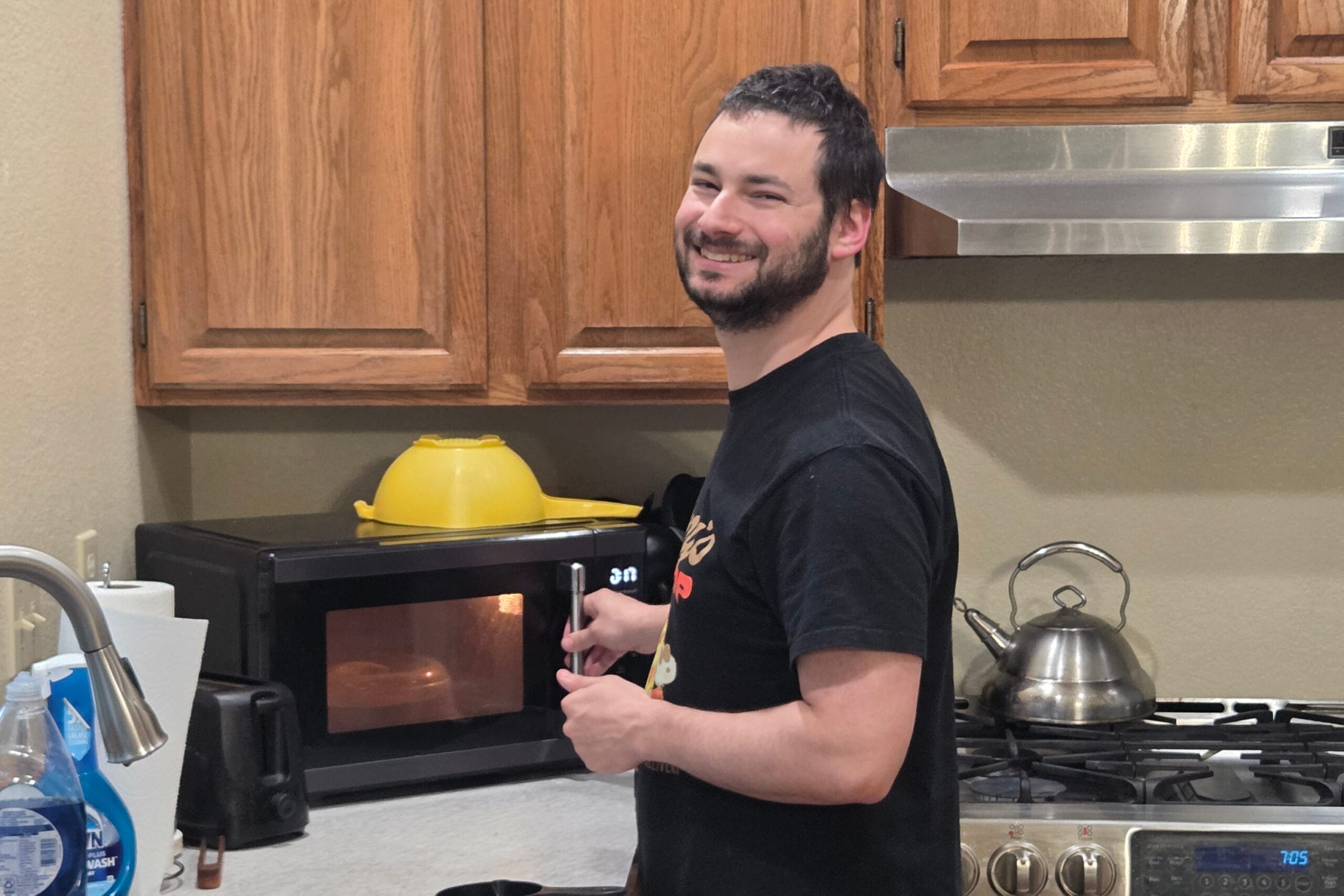 Smiling man in a black T-shirt standing in a kitchen beside a microwave, holding a utensil at chest height.
