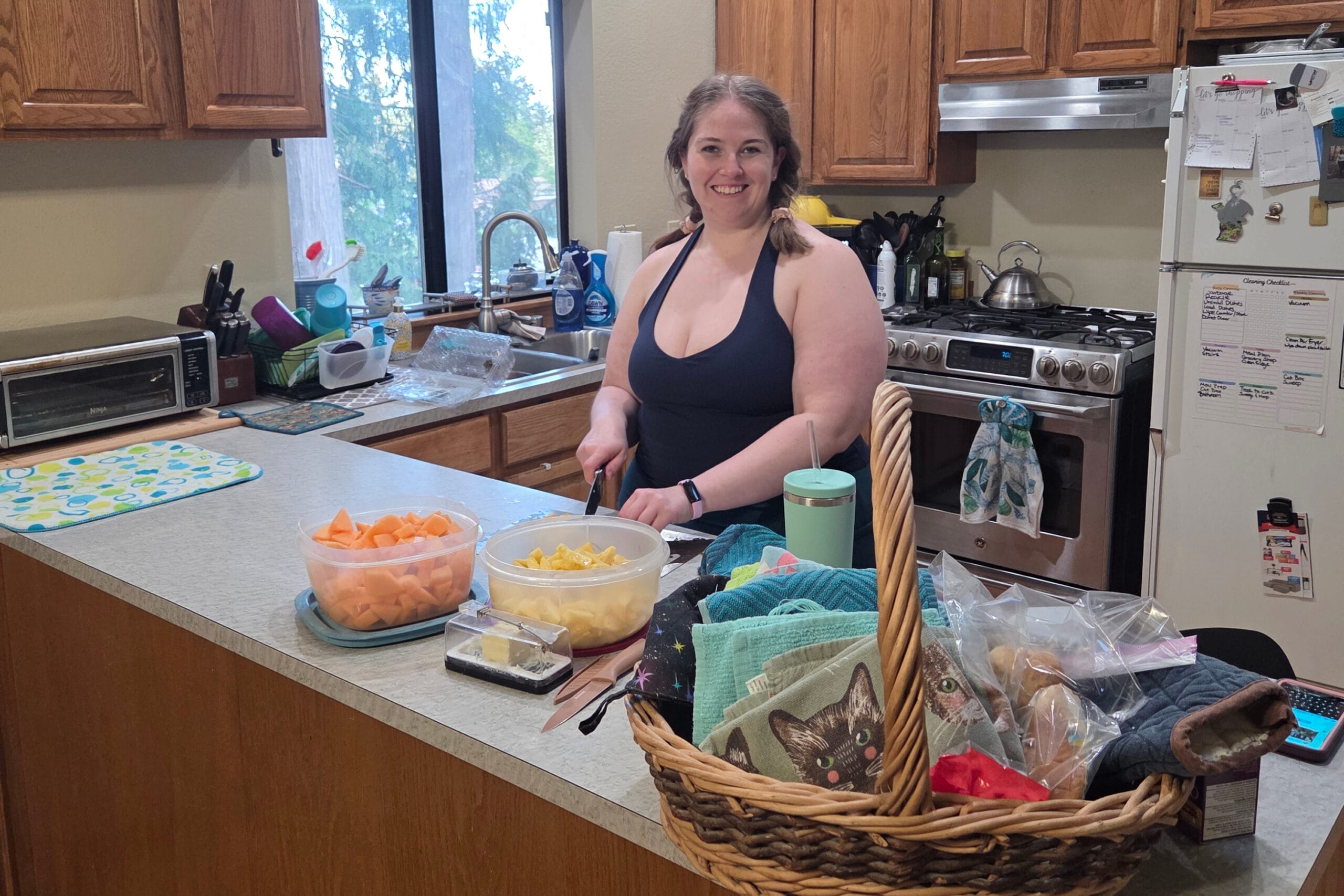 Smiling woman in a navy tank top chops fruit at a kitchen island; bowls of diced melon in the foreground.