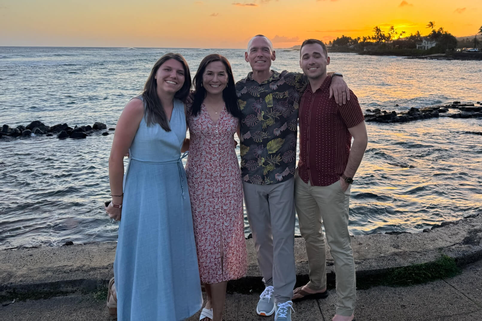 Four adults posing together on a seawall by the ocean at sunset, smiling with their arms around each other. The sea, rocky breakwater, and palm trees are in the background.