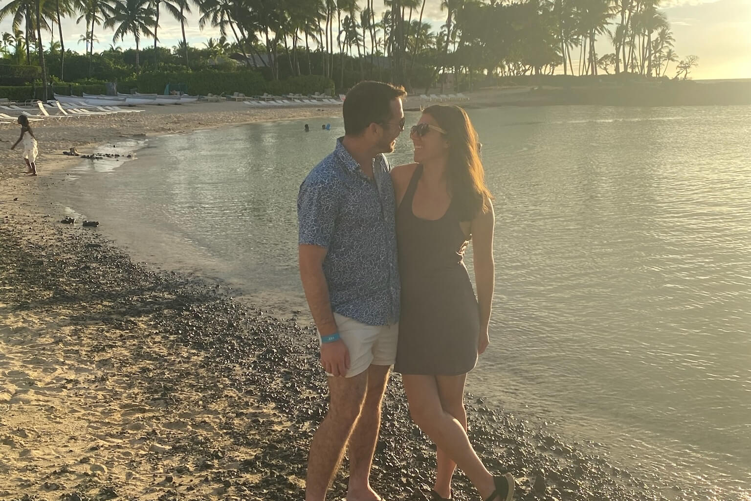 Couple standing close together on a sandy beach at sunset, smiling at each other with palm trees and calm water behind them.