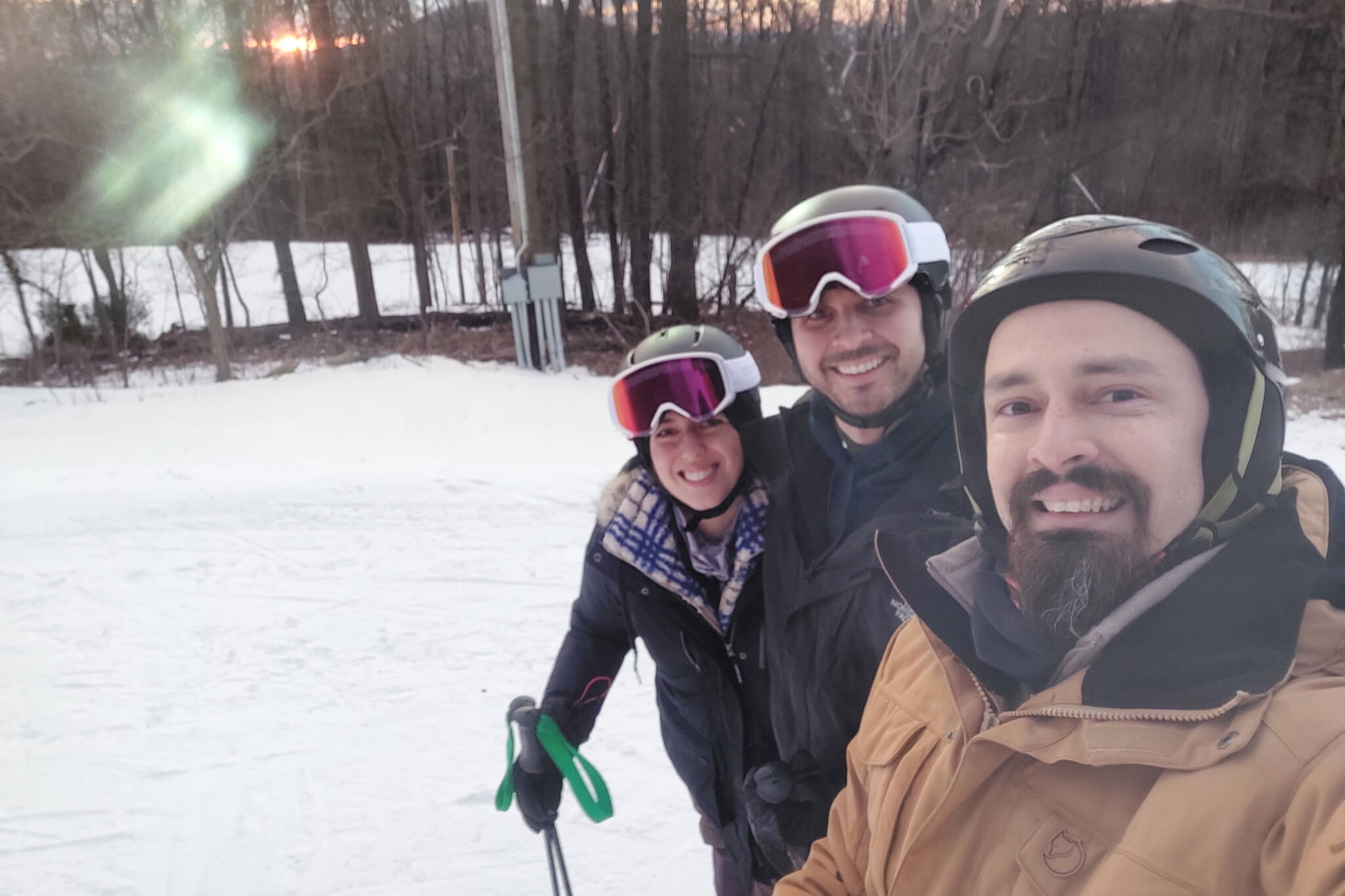 Three friends in ski gear pose for a selfie on a snowy day, wearing helmets and goggles with a forest background at sunset.