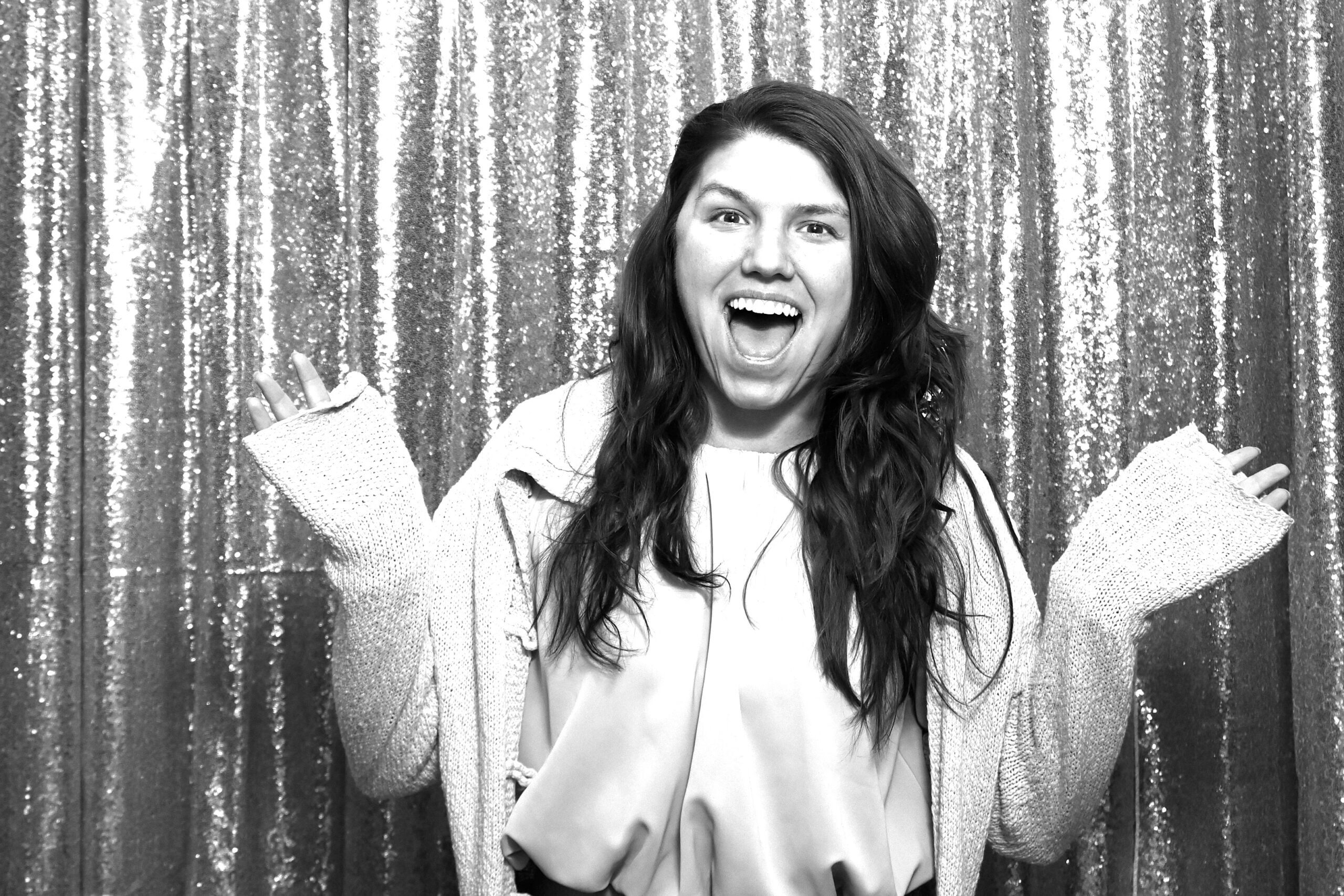 Black-and-white photo of a laughing woman with long hair and arms spread wide in front of a glittery backdrop.