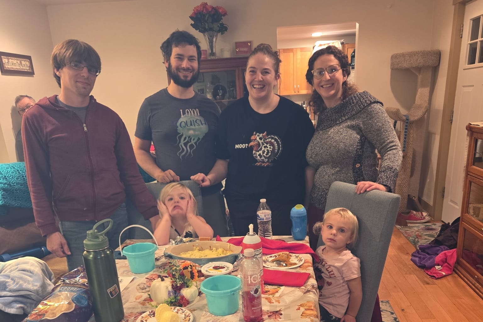 Group of five adults standing around a birthday or gathering table with kids seated, smiling in a cozy home dining room setting.