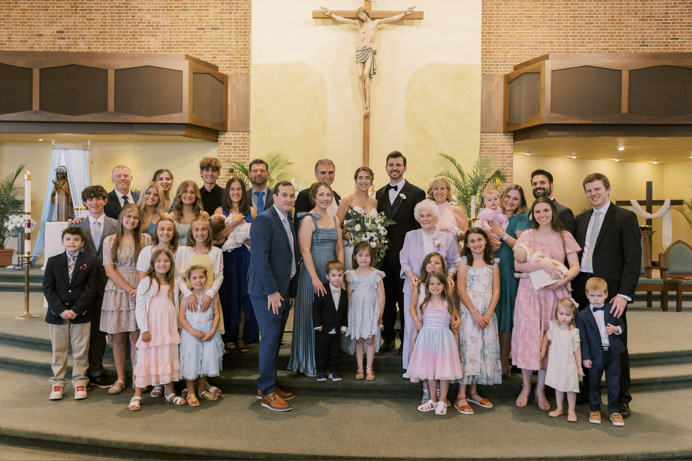Group of wedding guests in formal attire posing on church steps with a central crucifix behind them, including many children in pastel dresses and adults in suits and gowns.