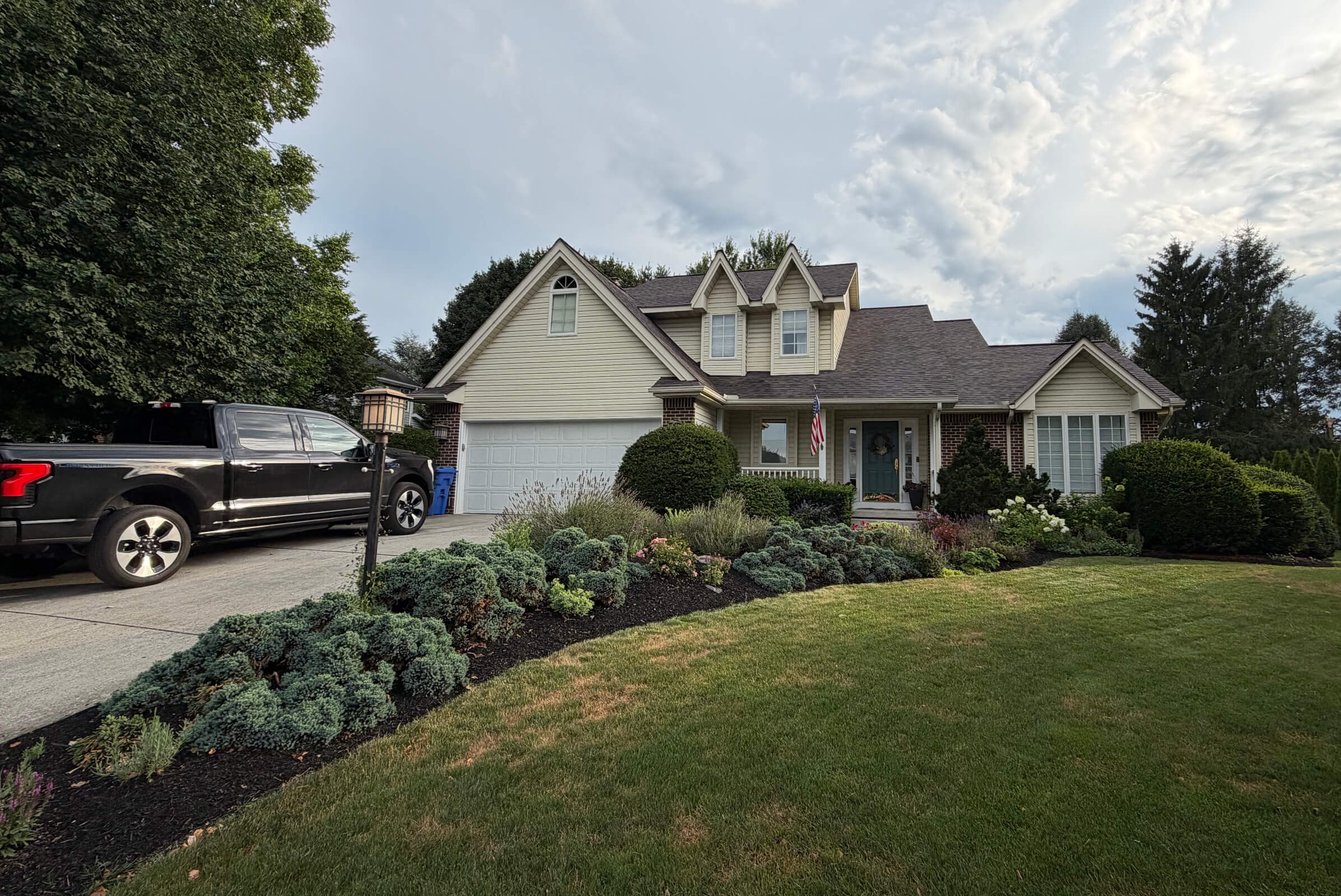 Suburban house with a two-car garage, well-kept front yard, and an American flag by the front door; black pickup in the driveway on the left.