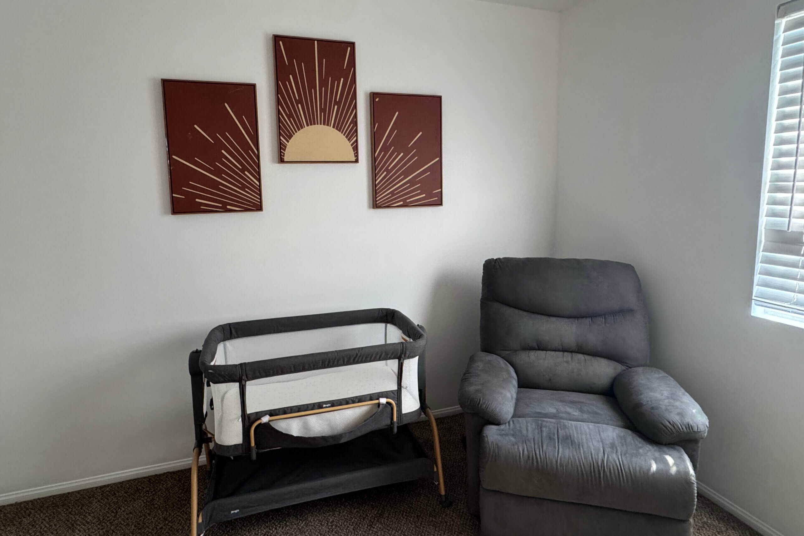 Gray recliner chair in a corner with a baby play yard on its left and a three-panel sunburst artwork on the wall above. window blinds visible to the right.