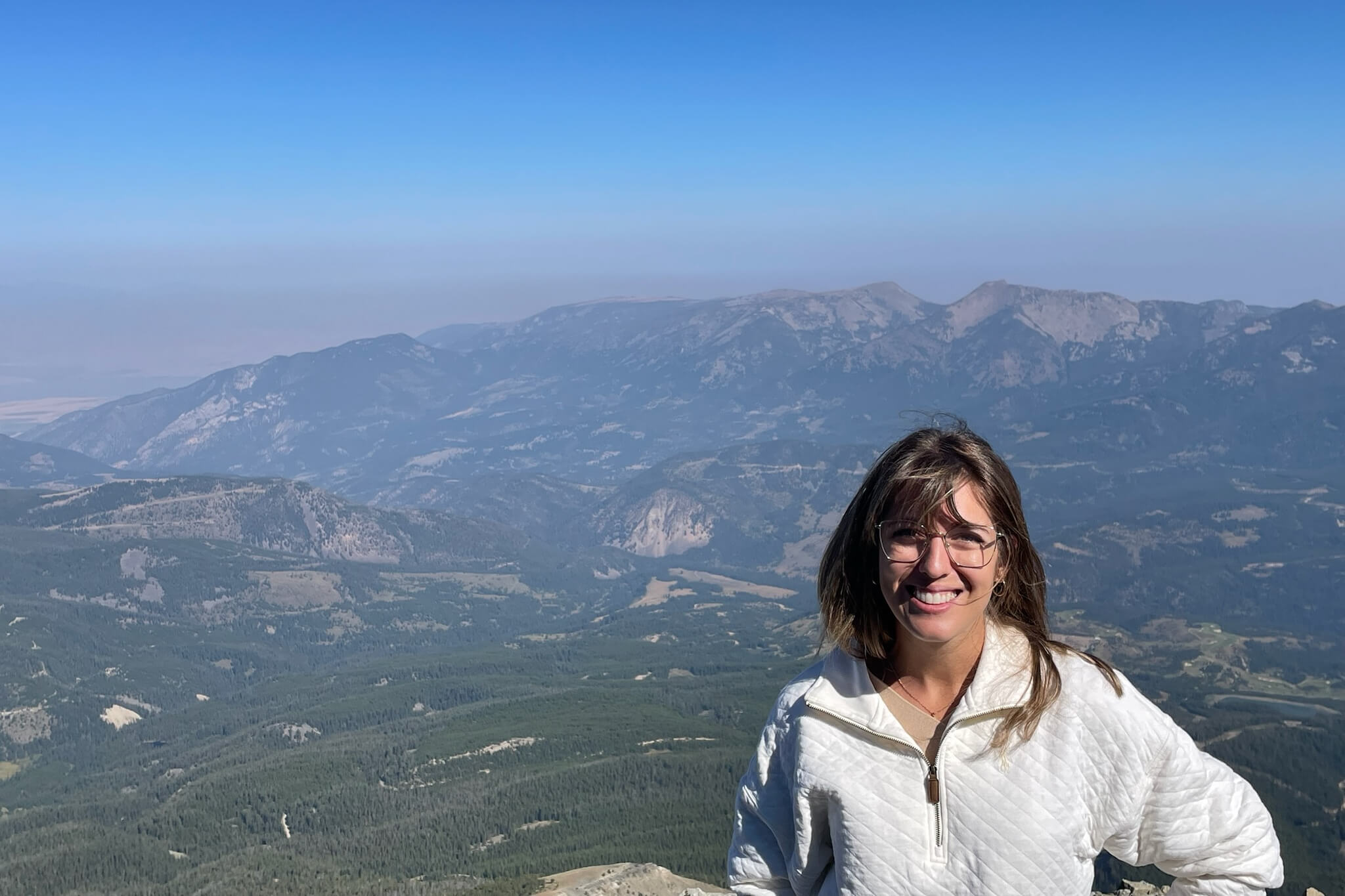 Smiling woman in a light jacket at a mountain overlook with blue sky and rugged peaks behind her.