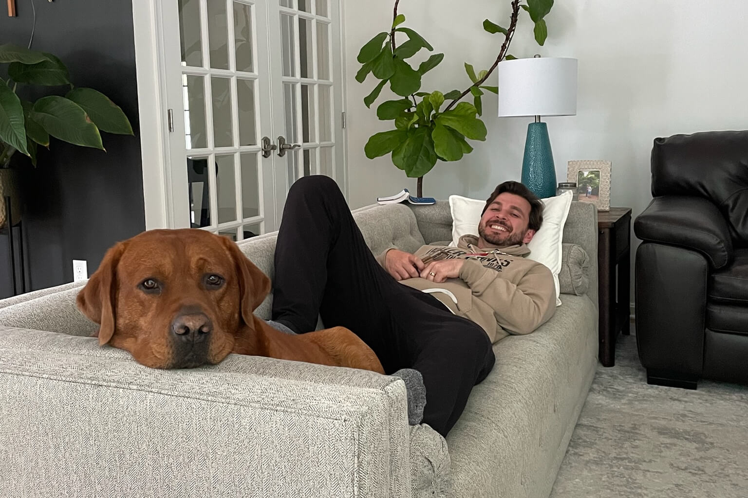Man reclining on a light gray sofa, smiling, with a large brown dog resting at the edge of the couch in a living room.