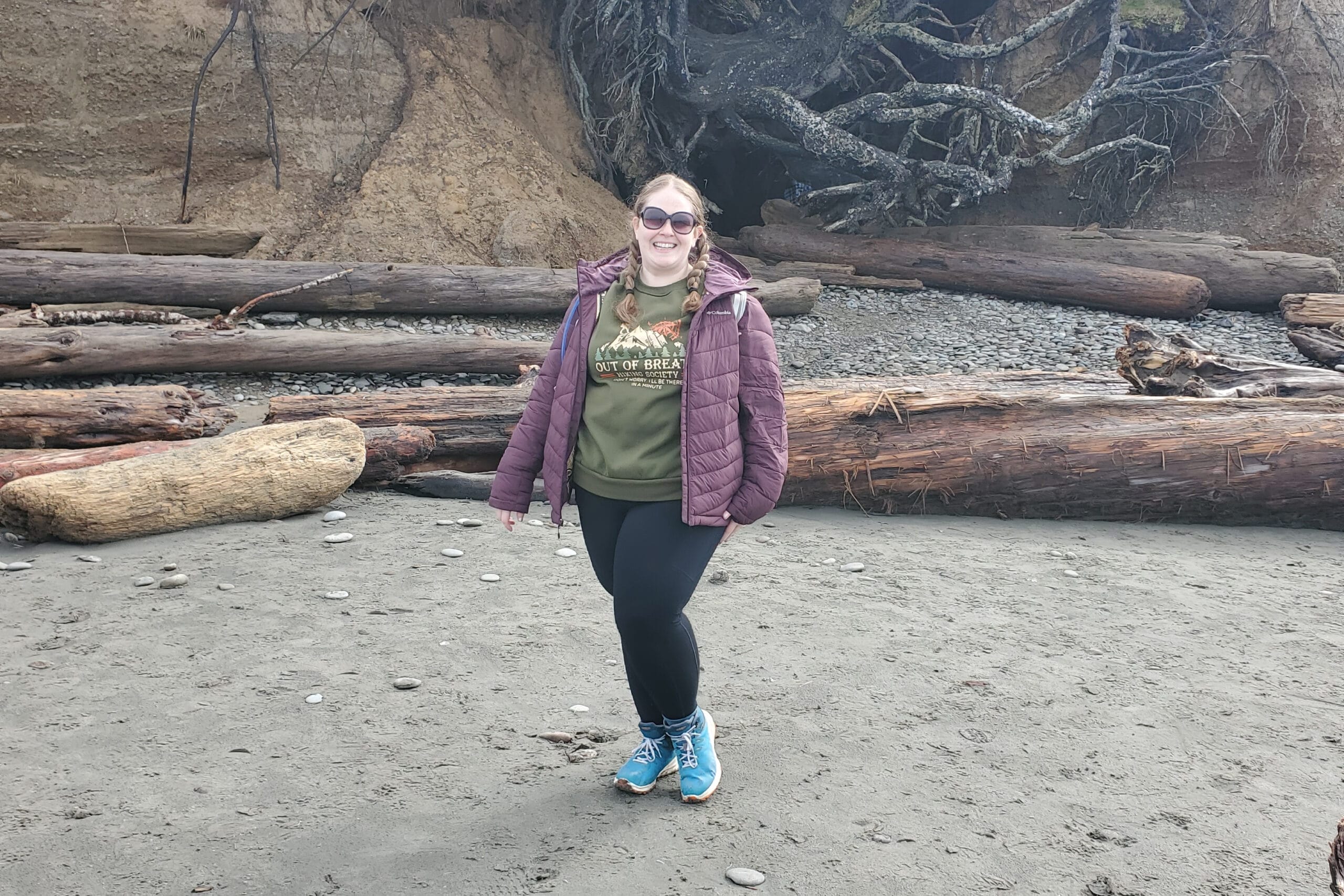 Smiling woman in a green shirt and maroon jacket stands on a sandy beach with large driftwood logs behind her and a rocky cliff.