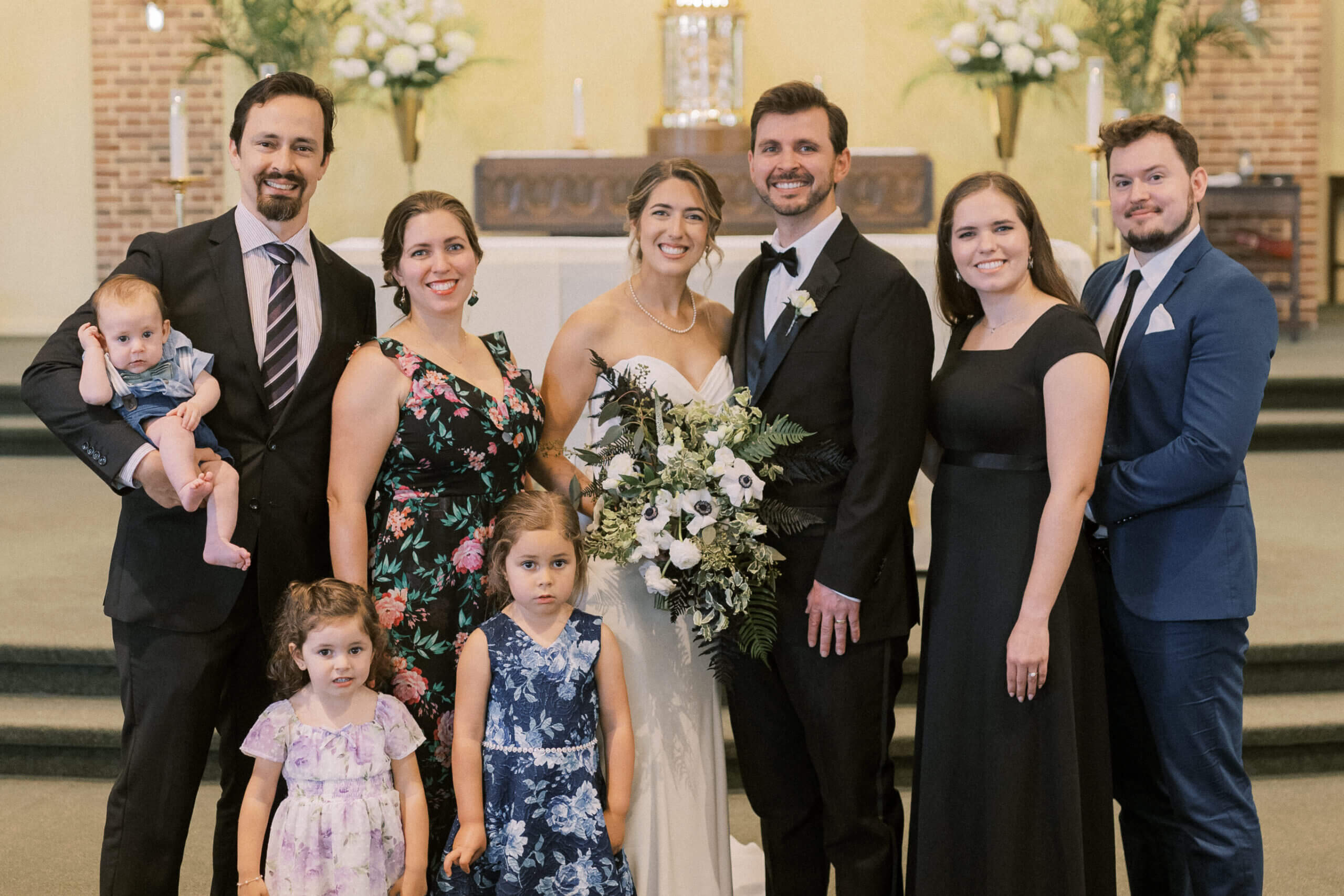 Bride and groom pose with bridal party in a church, flanked by bridesmaids and groomsmen, plus two young girls in front and a man holding a baby on the left.
