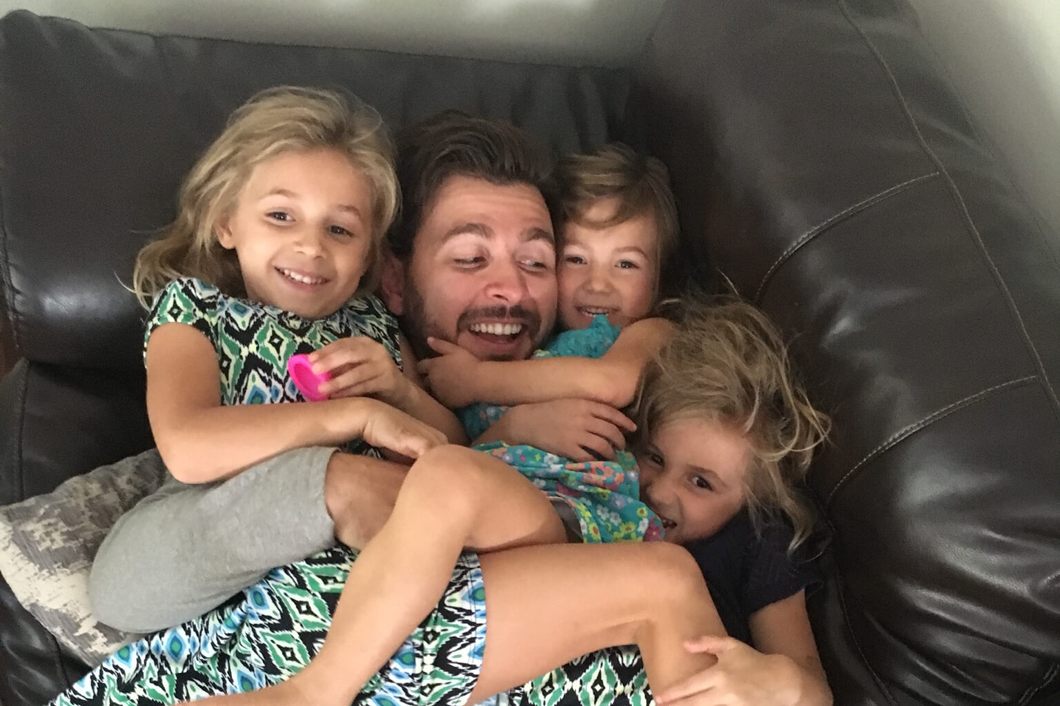 Dad is hugged by four smiling children on a dark leather couch, sharing a cozy family moment.