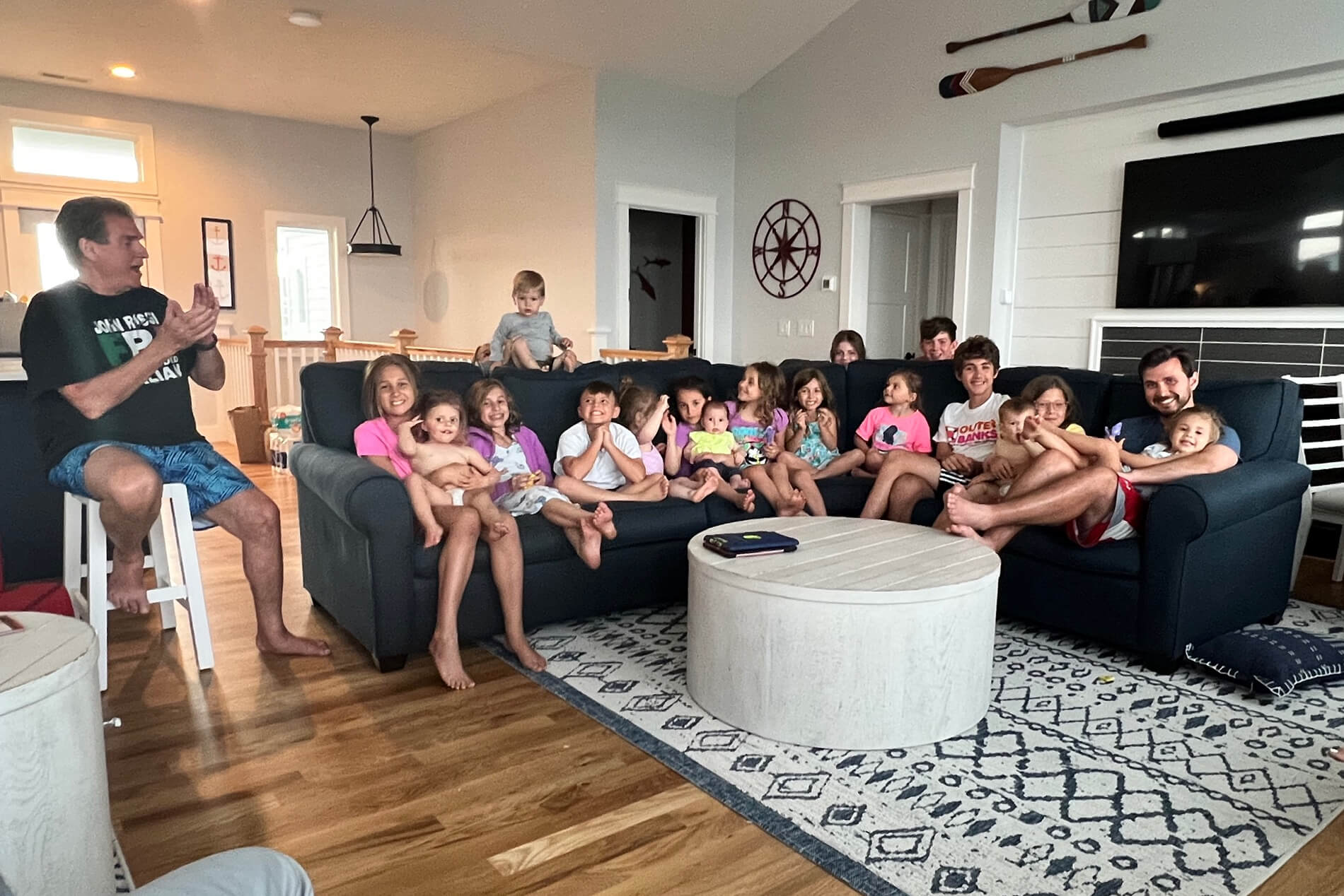 Family group photo in a bright living room: many children sit on a dark blue sectional while a man on a stool claps nearby.