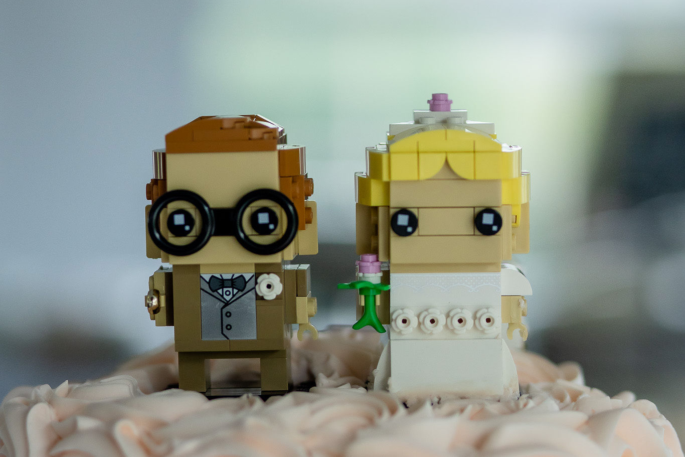 Two LEGO wedding figures, a groom with glasses and a bow tie and a bride in a white gown, stand on a pink-petal cake topper
