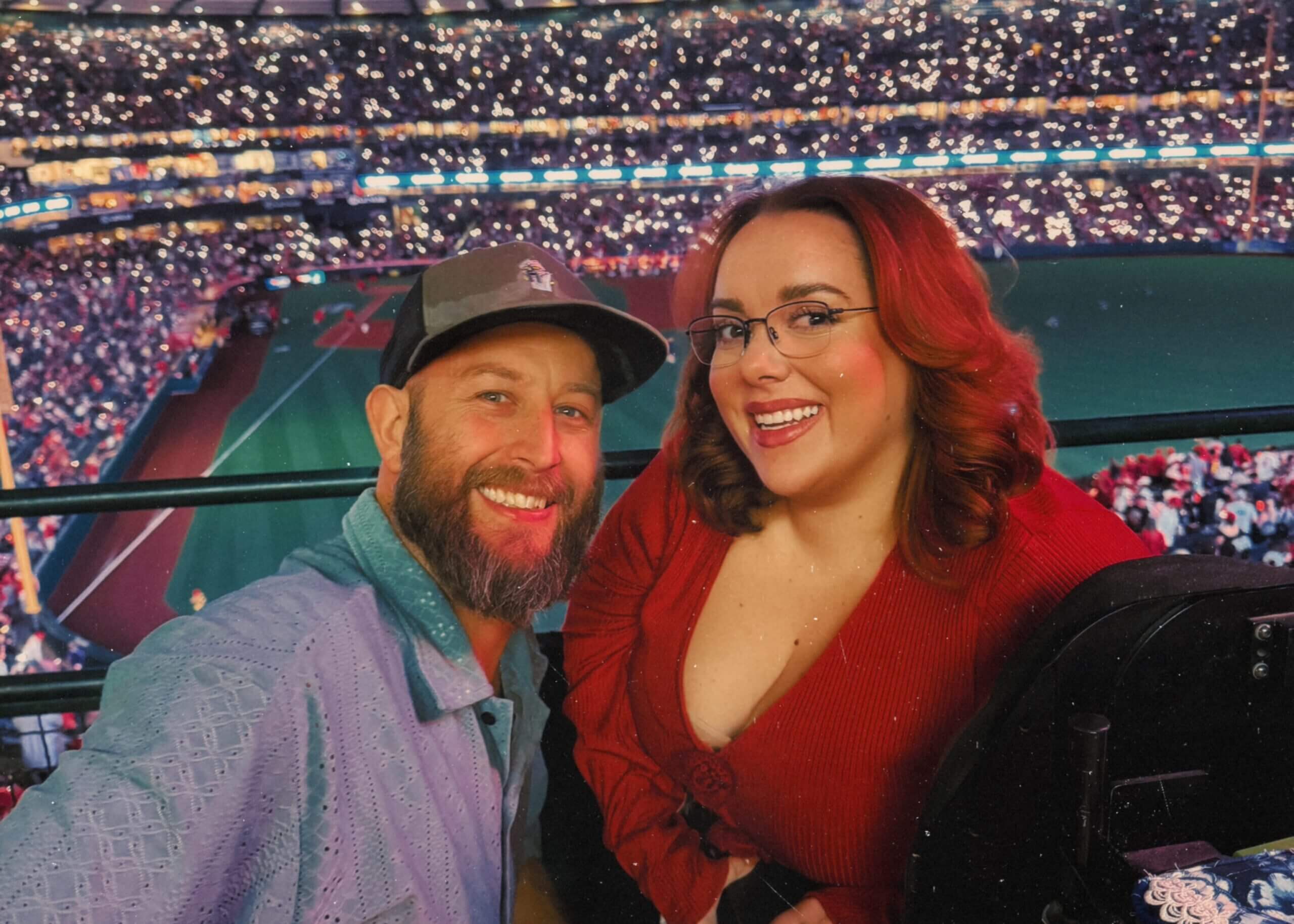 A smiling couple taking a selfie at a baseball stadium during a night game, with a crowd and bright stadium lights behind them.