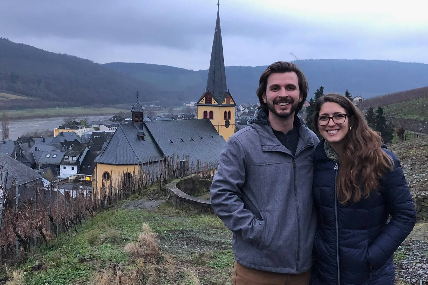 Smiling couple in jackets pose on a hillside with a yellow church and tall spire behind them in a valley village on a cloudy day.