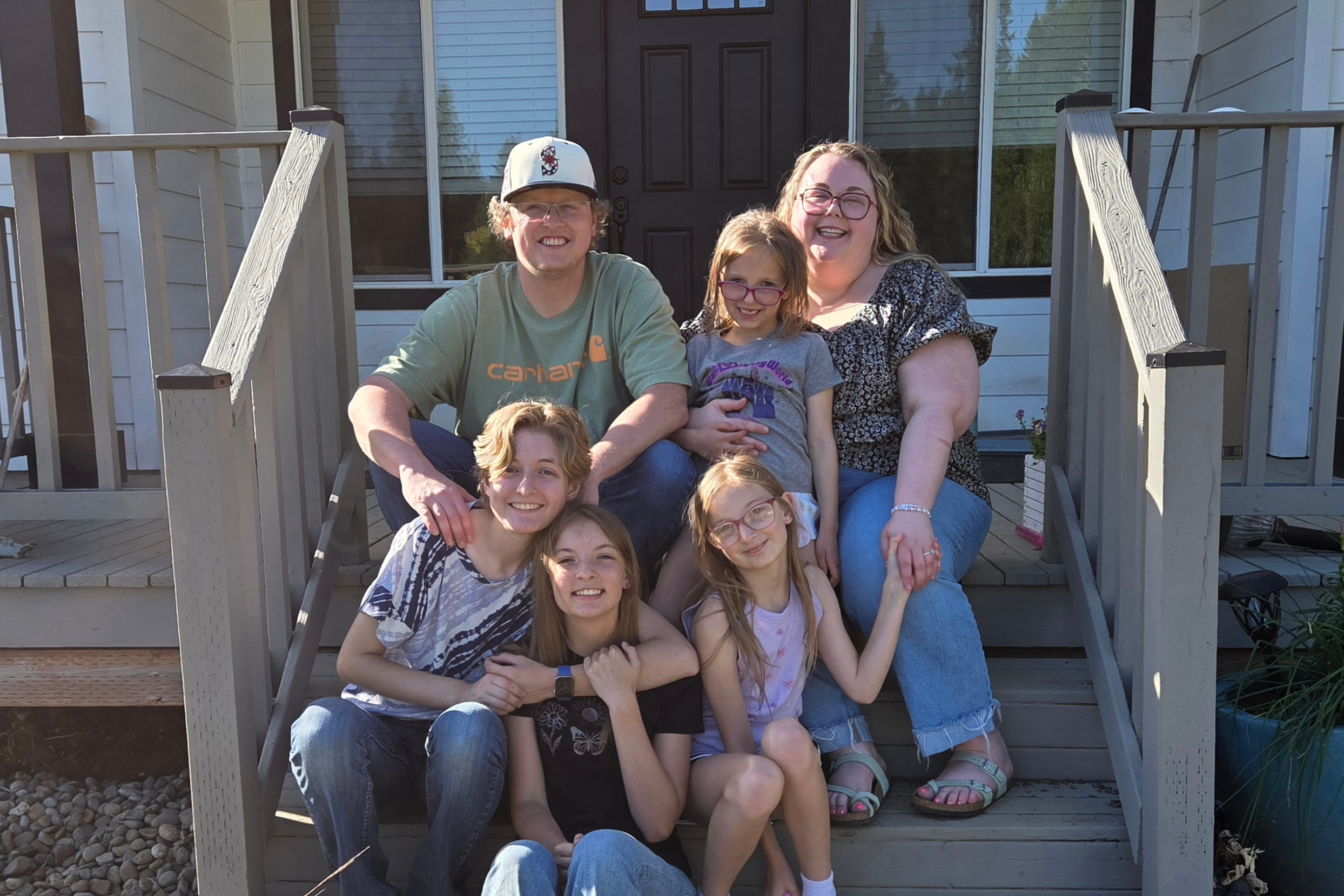 Family of six posing on front porch steps, smiling at the camera with a house and railing behind them.