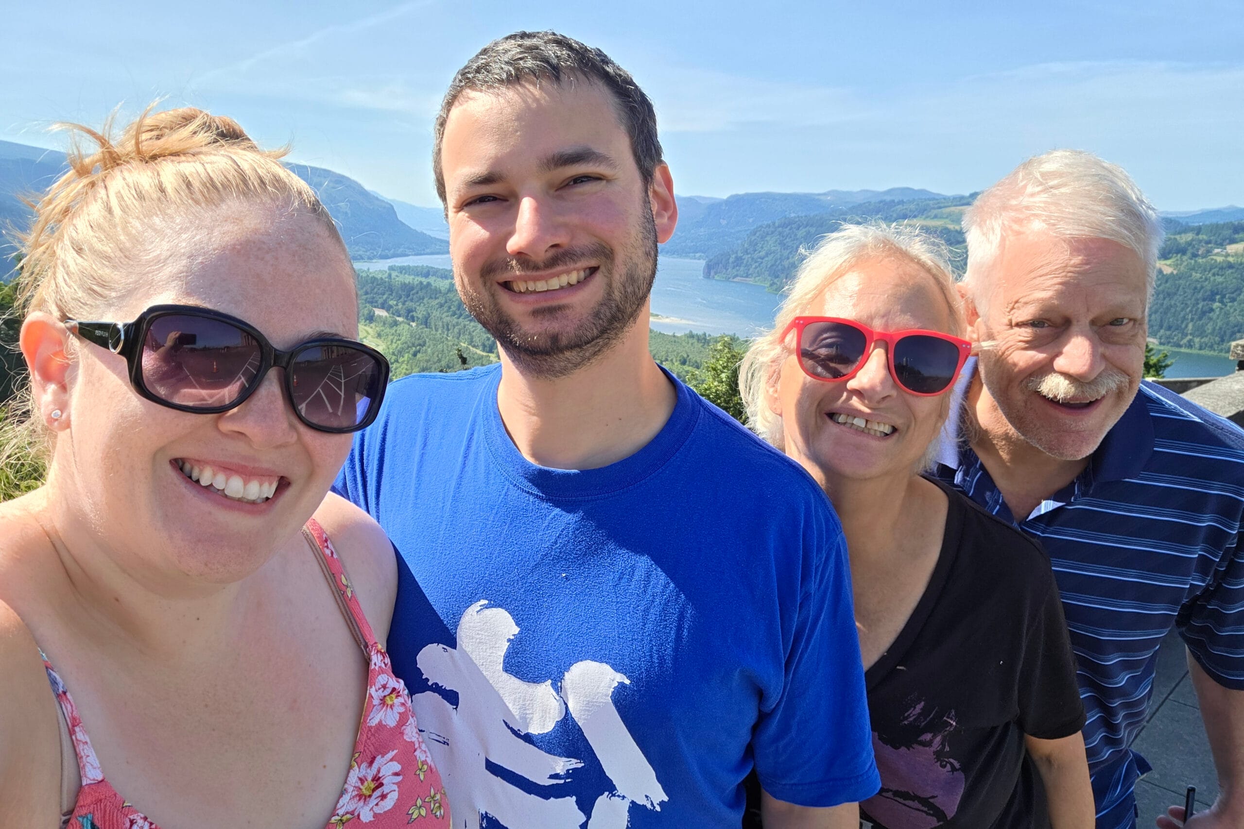 Four friends taking a sunny selfie with a lakeside and green hills in the background, smiling at the camera.