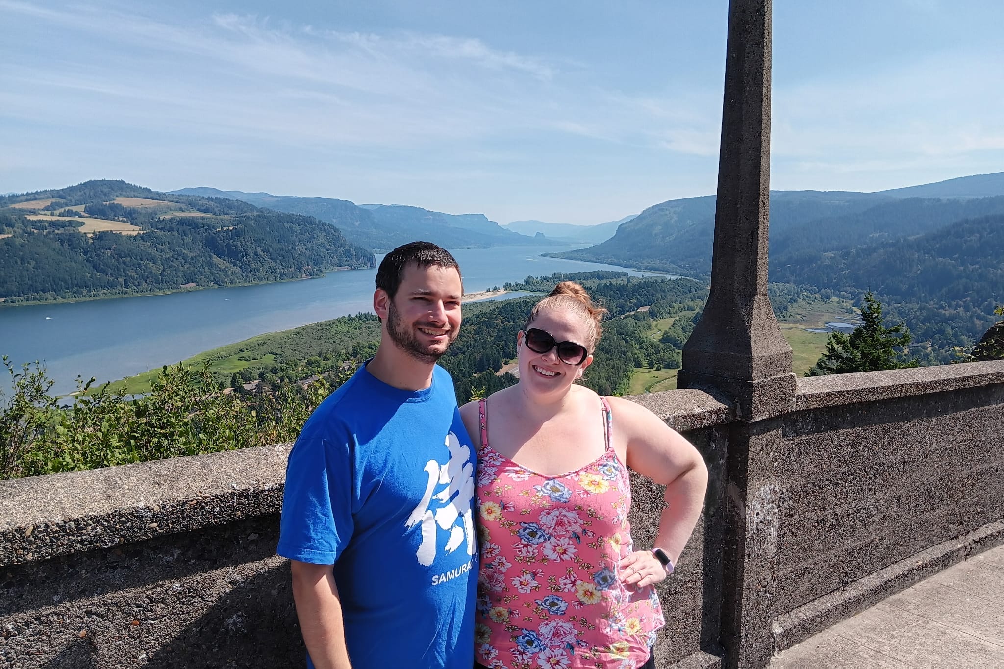 Smiling couple posing at a stone overlook with a river valley and green hills in the background.
