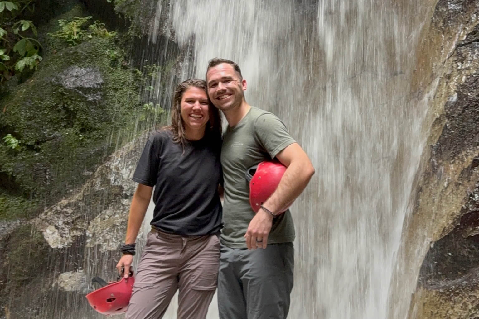 Couple posing and smiling in front of a waterfall, wearing outdoor gear and holding red helmets for rafting or climbing.
