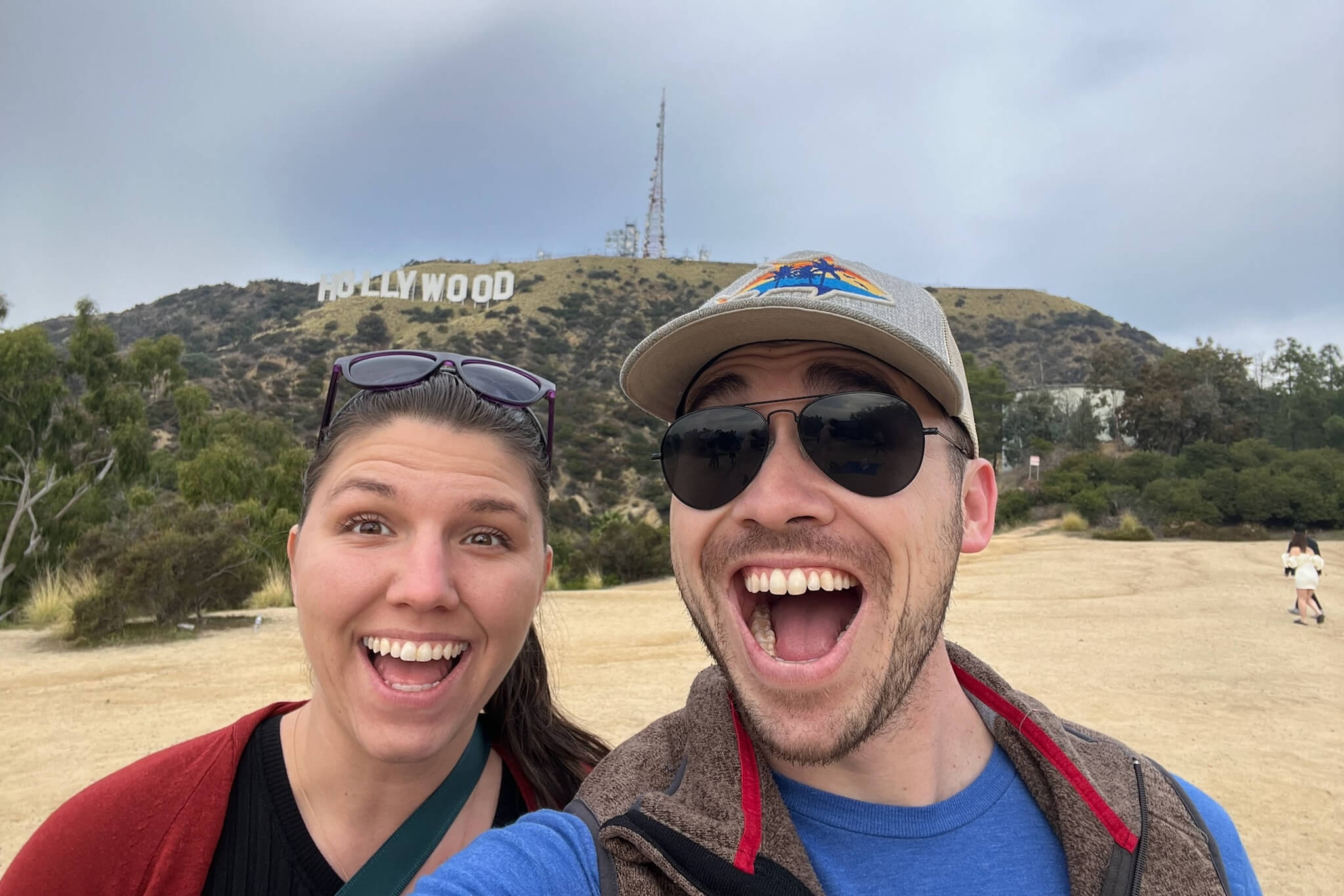 Two friends take a cheerful selfie with the Hollywood sign on a hill in the background in clear daylight.