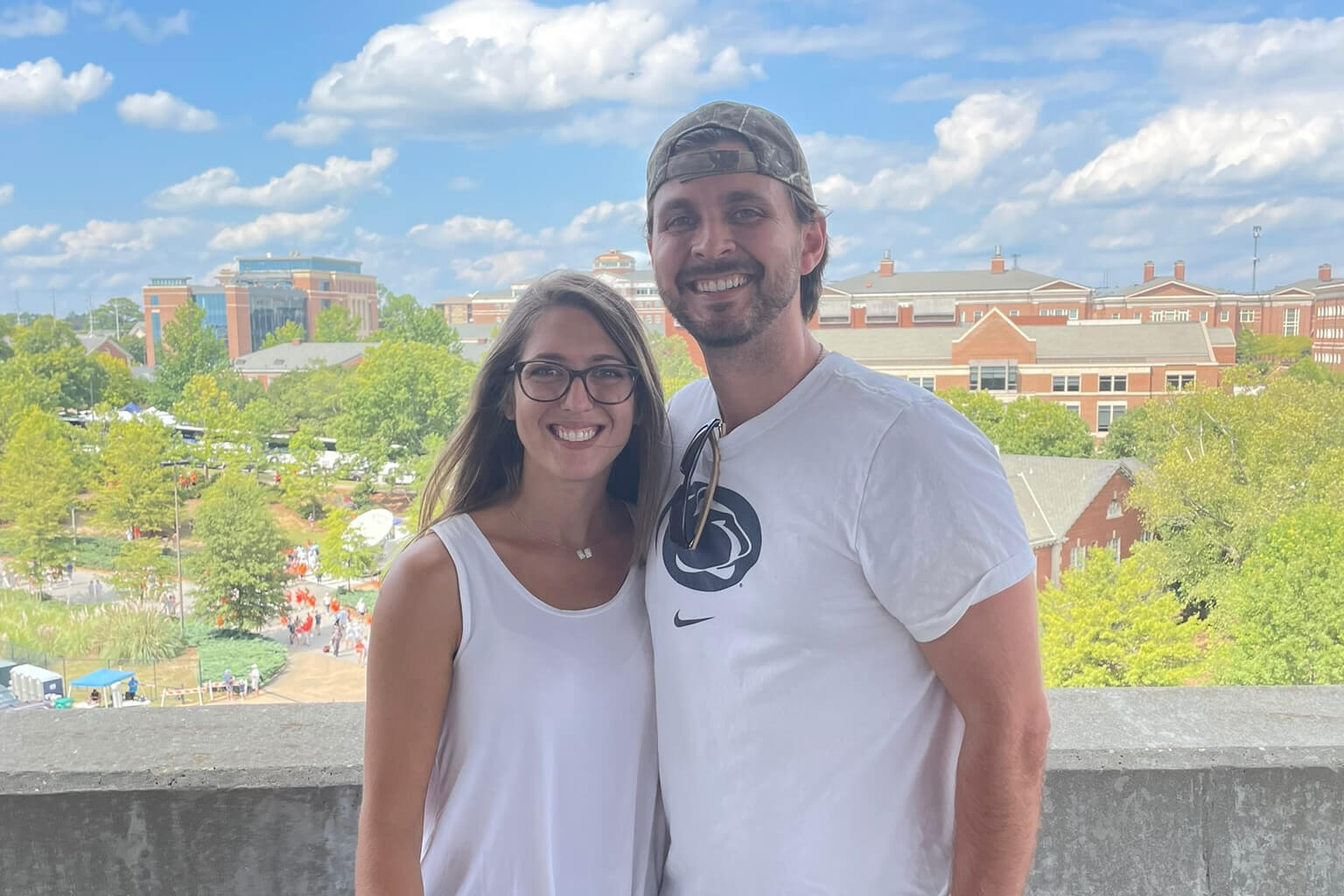 Smiling couple standing arm-in-arm on a balcony with a campus skyline, trees, and a blue sky in the background.
