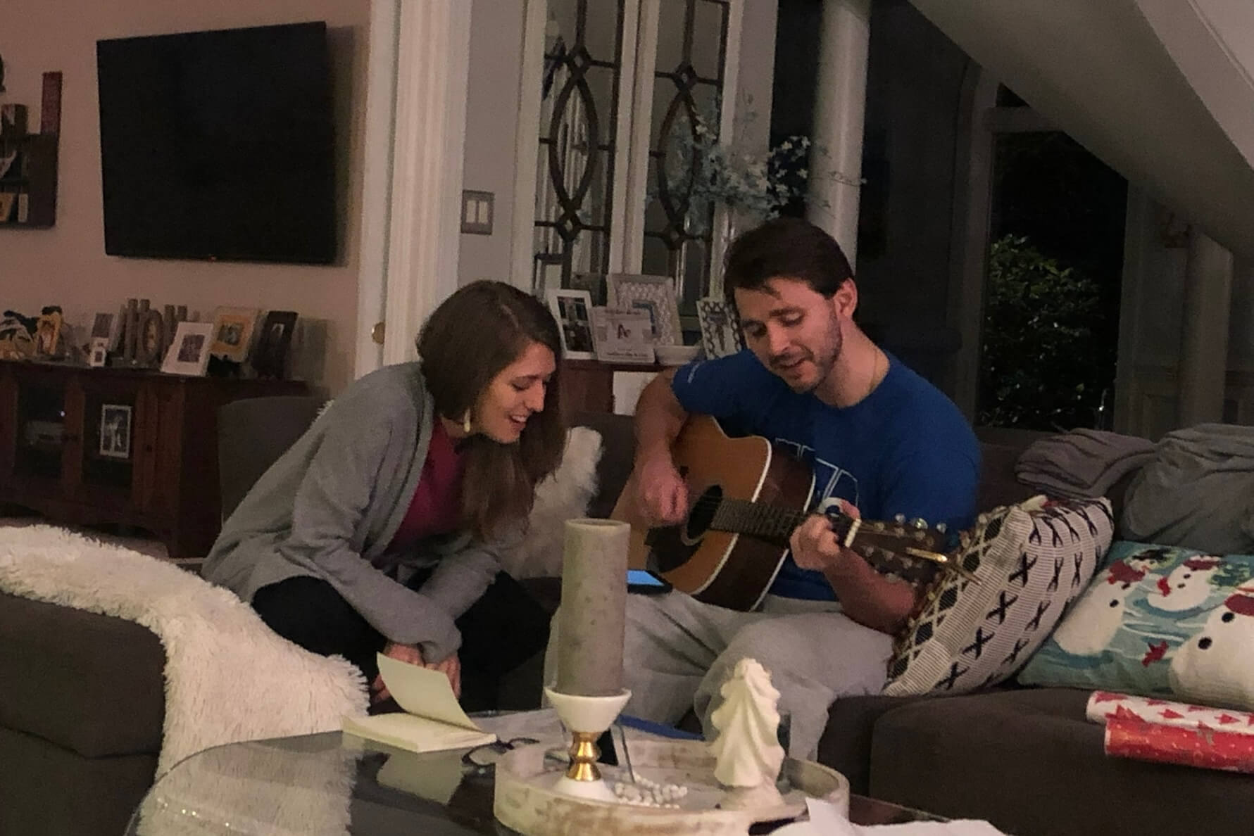 Two friends on a sofa: one man strums an acoustic guitar while a woman reads from a note nearby in a cozy living room setting.