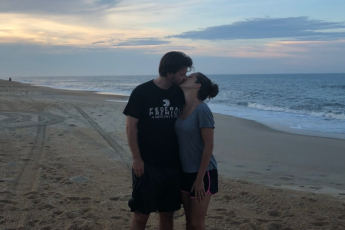 Couple kissing on a sandy beach at sunset with the ocean in the background.