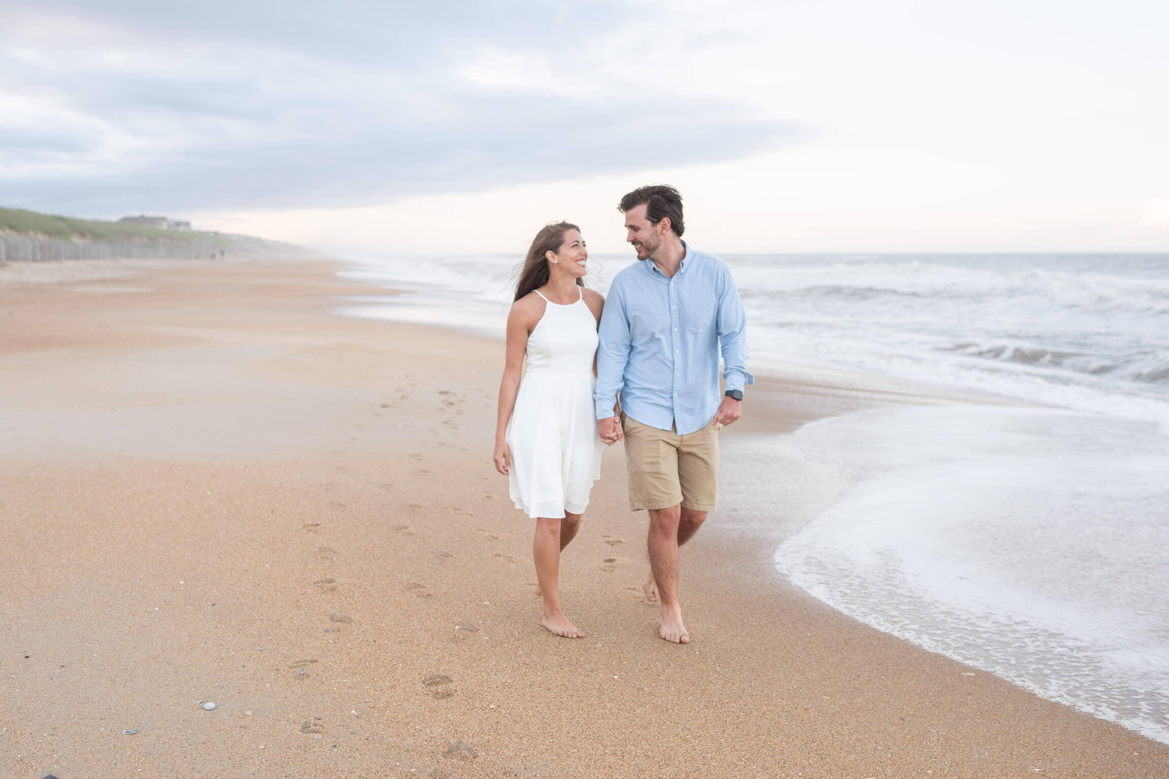 Couple walking barefoot on the beach, holding hands and smiling as waves wash in.