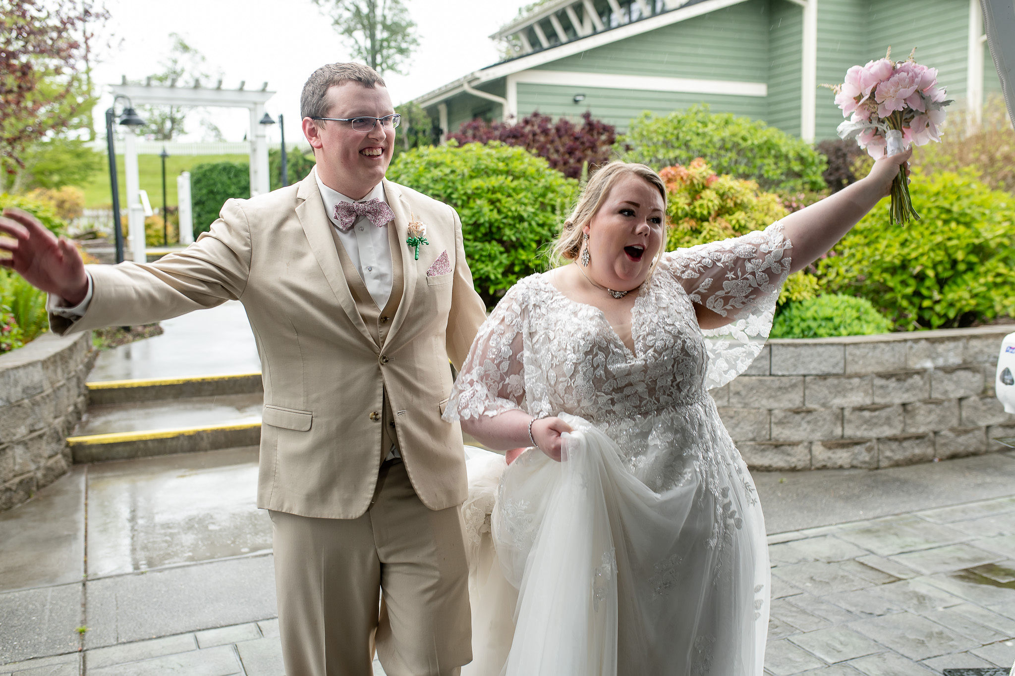 Joyful newlyweds outdoors, she in a lace wedding gown with a pink bouquet, he in a tan suit and bow tie, cheering with arms raised.