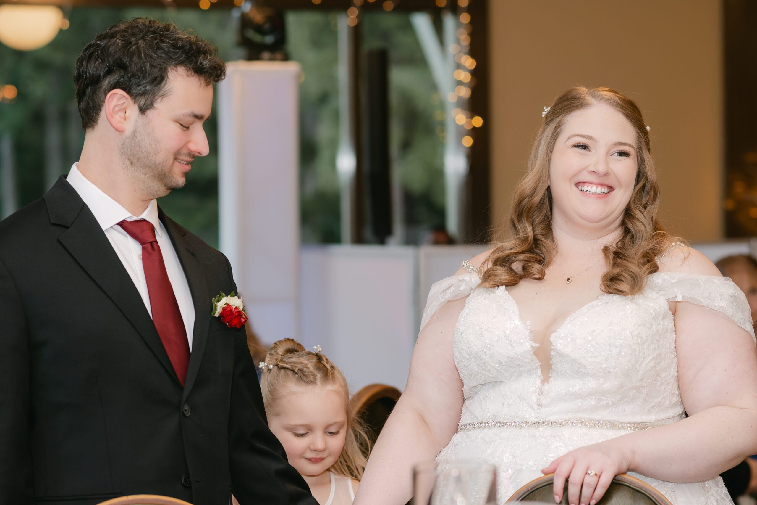 Bride in a white gown and groom in a black suit smile during a wedding, with a young girl between them.