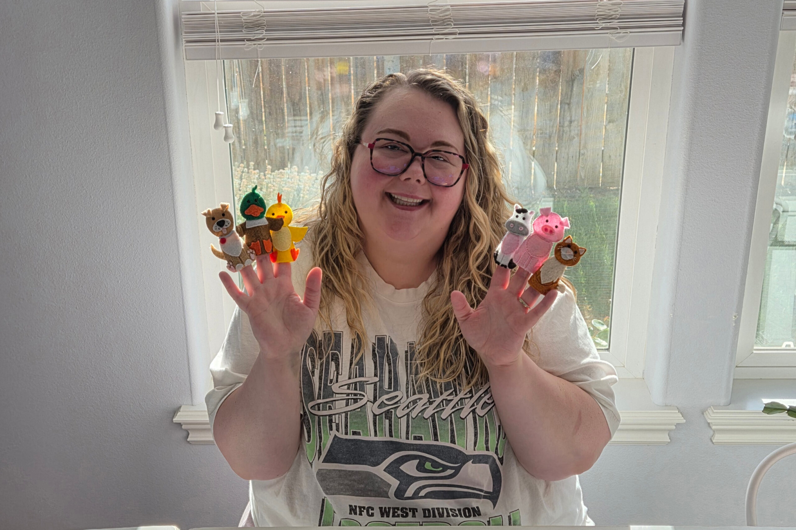Smiling woman with glasses holding colorful felt animal finger puppets in front of a window.