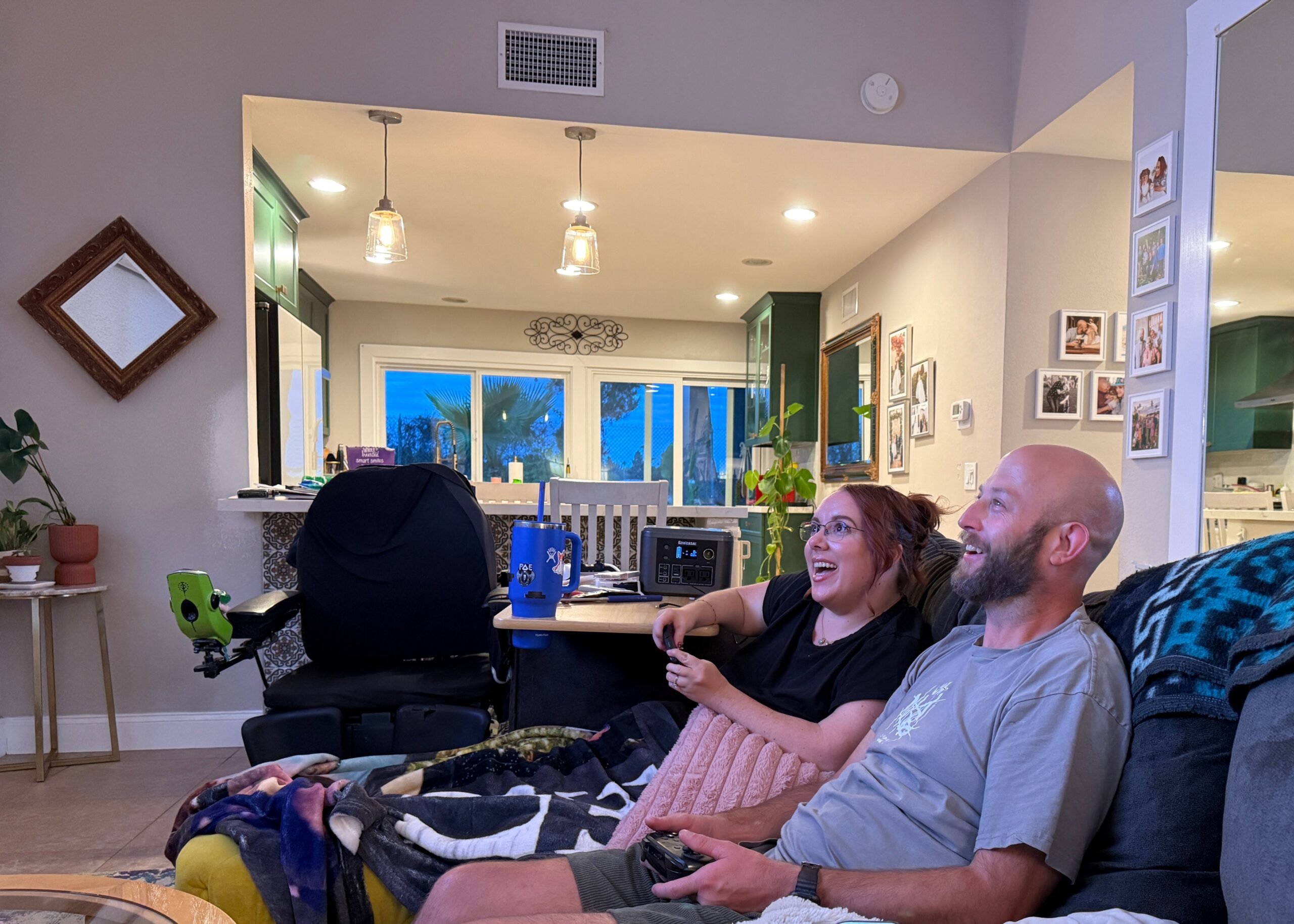 Group of people relaxing on a couch in a bright, open living room, smiling and watching something off-camera; a wheelchair is visible on the left beside the table of supplies.