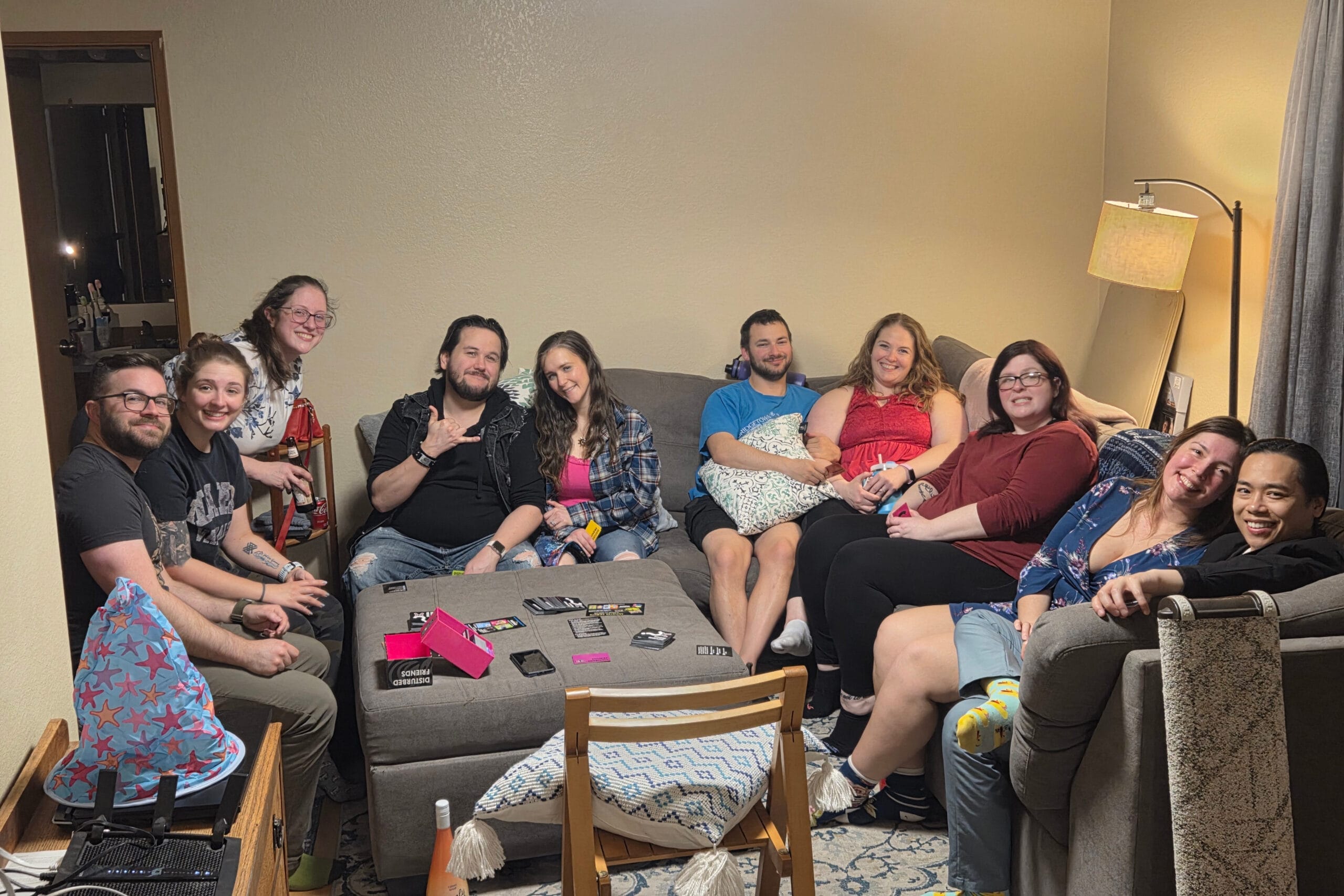 A group of friends sitting on a living room sofa, smiling at the camera with a table of game cards in front.