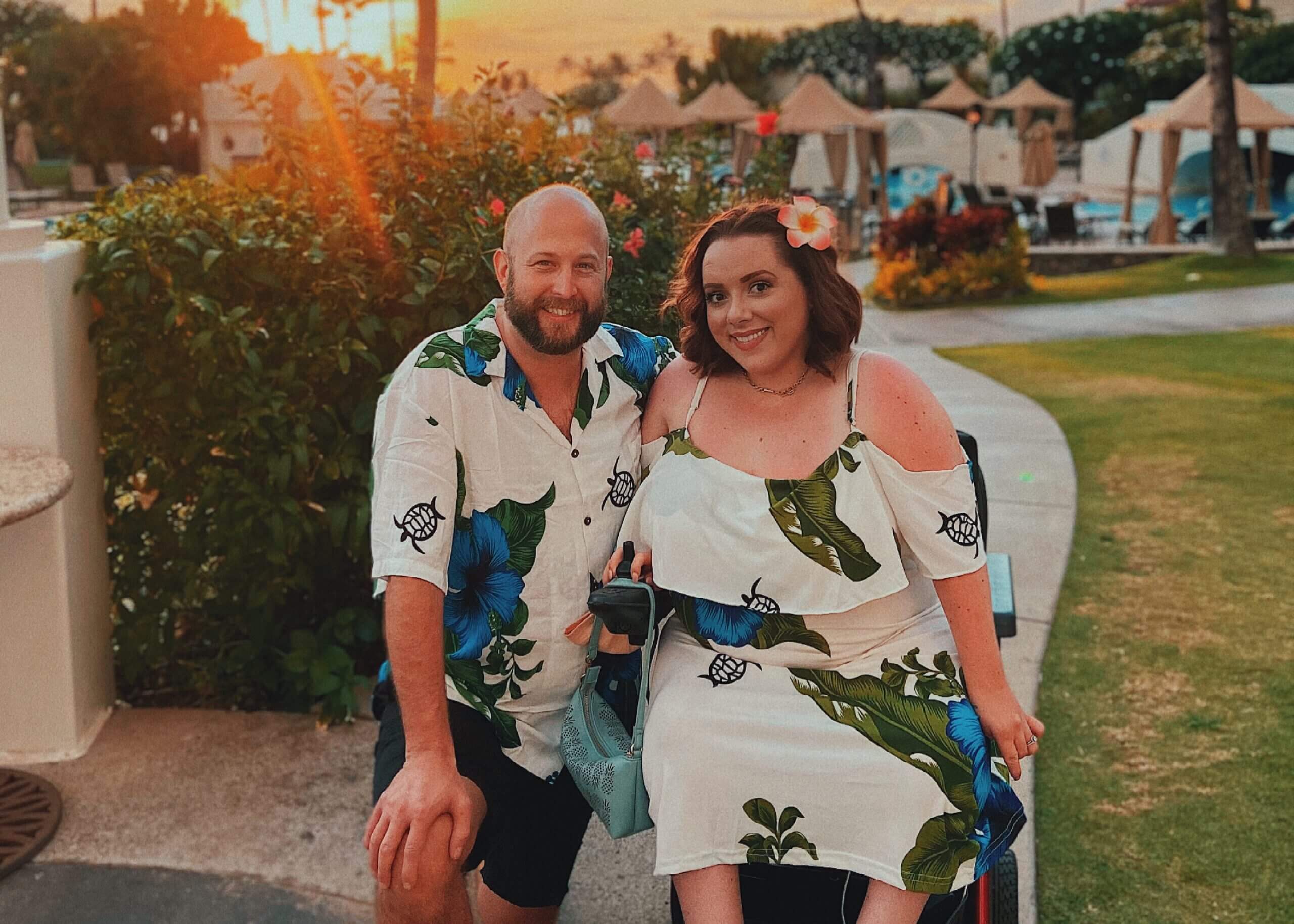 Smiling couple in tropical print outfits outdoors at a resort with a sunset glow.