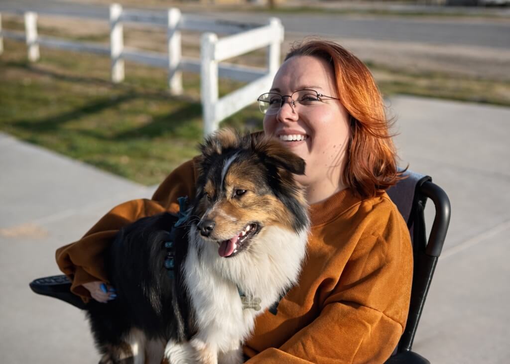 Smiling woman in a wheelchair holds a fluffy black-and-white dog outdoors near a white fence.