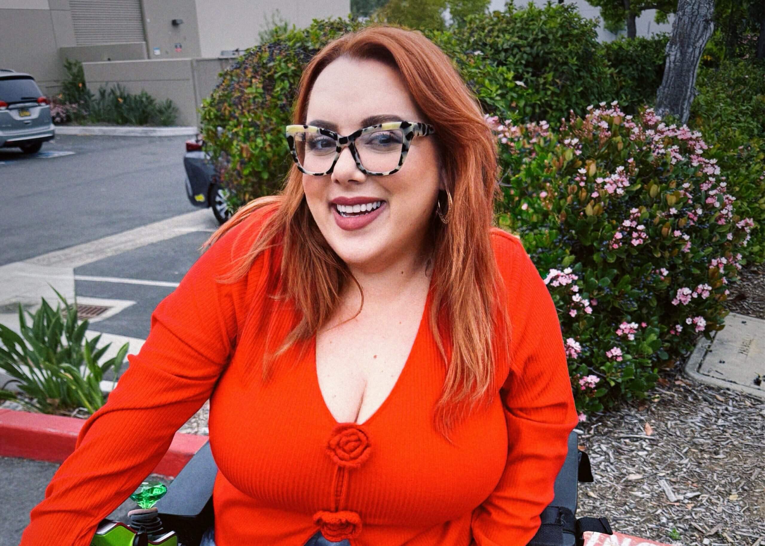 Smiling woman with red hair and patterned glasses in an orange sweater outdoors near flowering shrubs in a parking lot.