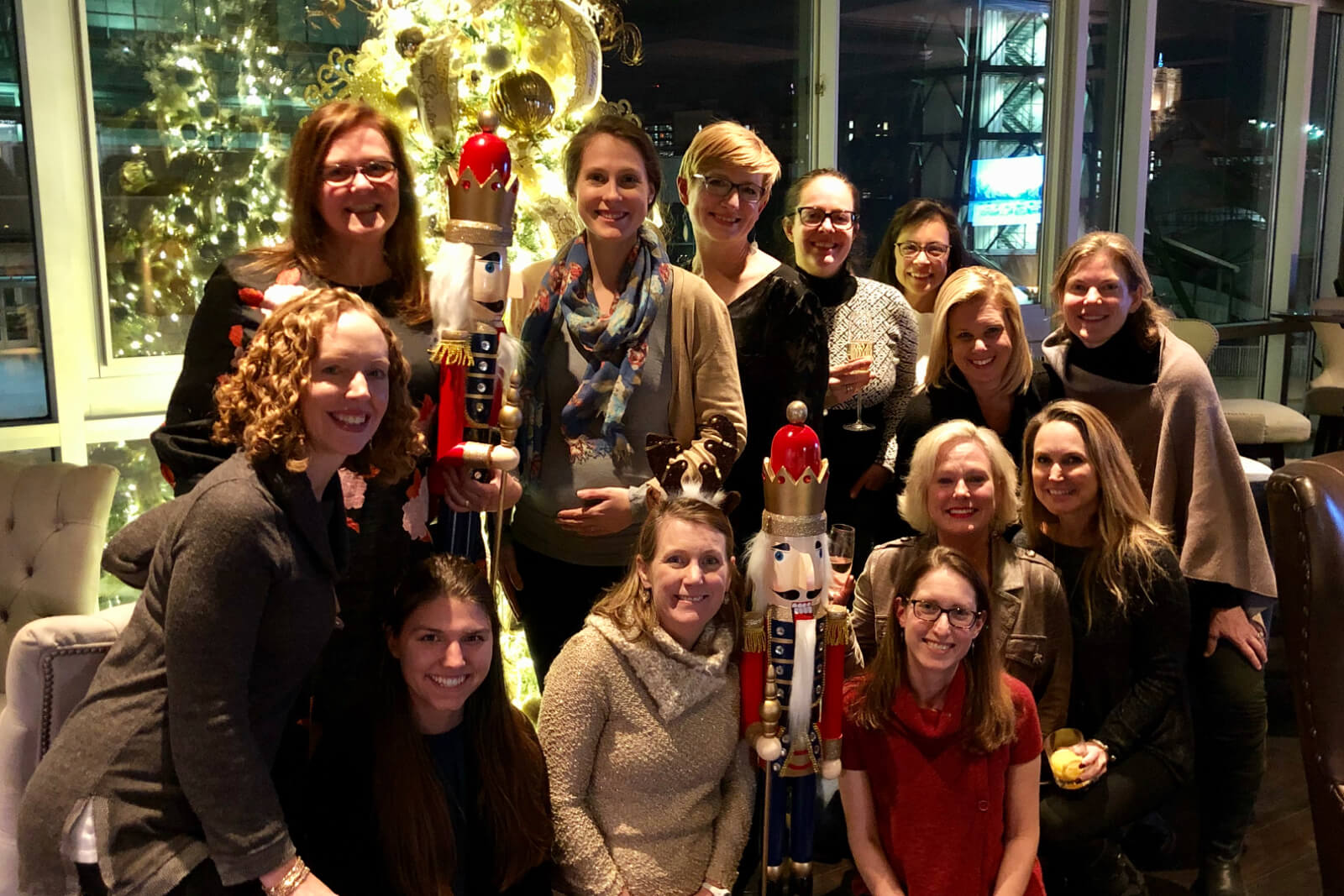 Group of women posing indoors with oversized nutcracker figures and a decorated Christmas tree in the background, smiling at the camera.